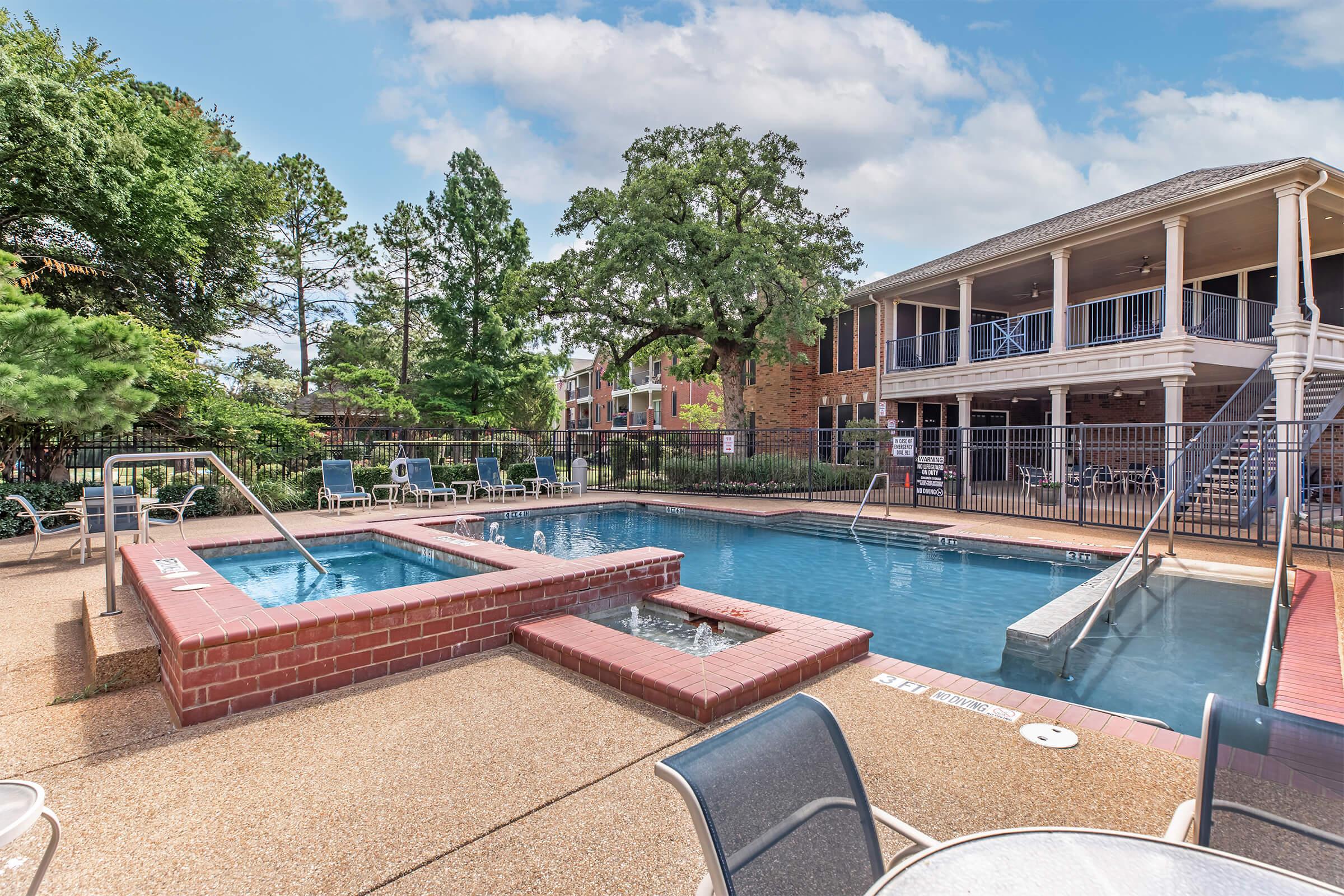 A swimming pool area featuring two pools, one larger and a smaller hot tub, surrounded by lounge chairs. Lush trees and greenery provide a natural backdrop. Adjacent to the pool area is a two-story building with a covered porch, creating a relaxing atmosphere. The sky is clear with a few clouds.
