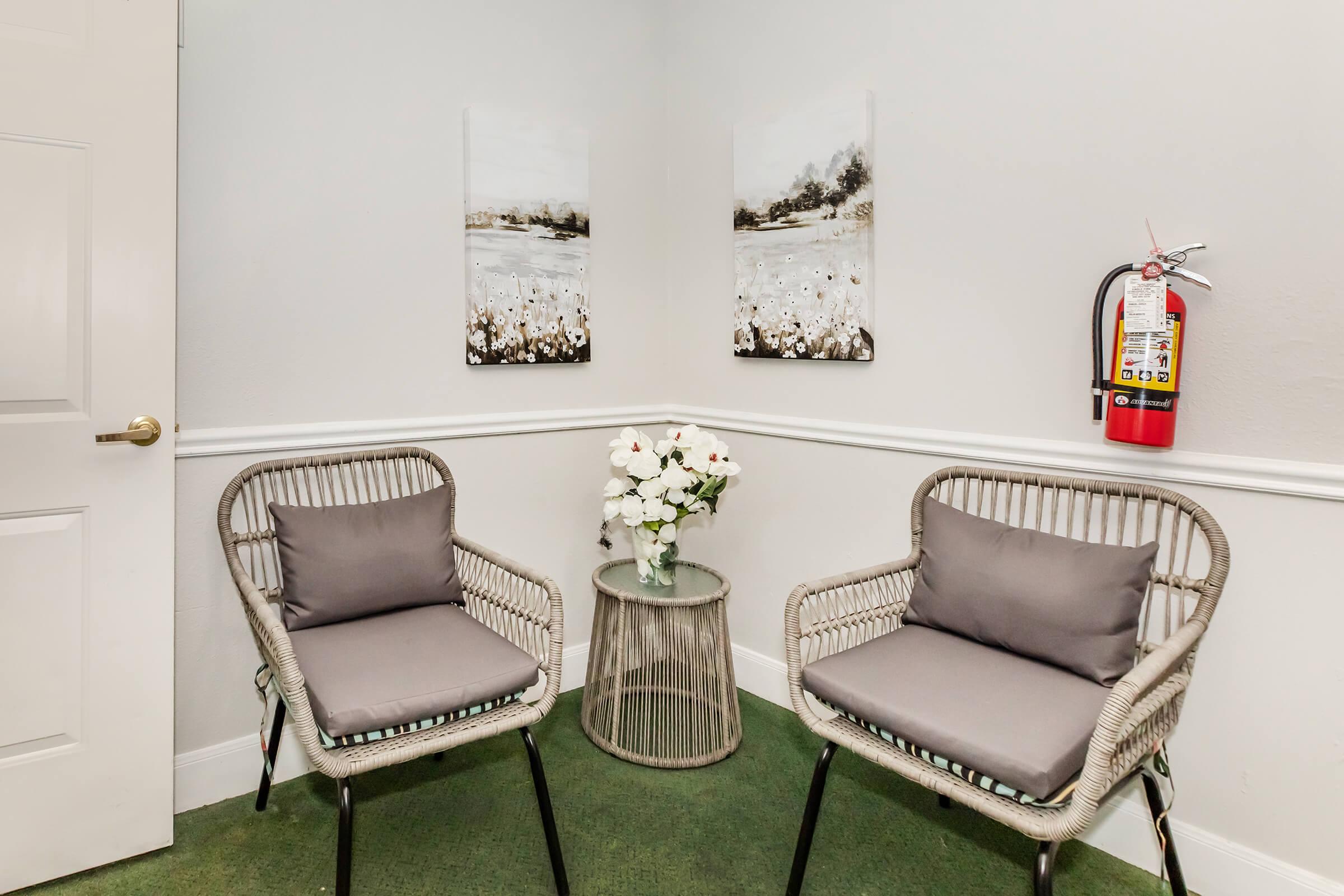 A cozy corner seating area featuring two wicker chairs with gray cushions, a small round table in the center, and a vase of white flowers. The walls are adorned with two abstract landscape paintings, and a fire extinguisher is mounted nearby. The floor has green carpeting, creating a welcoming atmosphere.