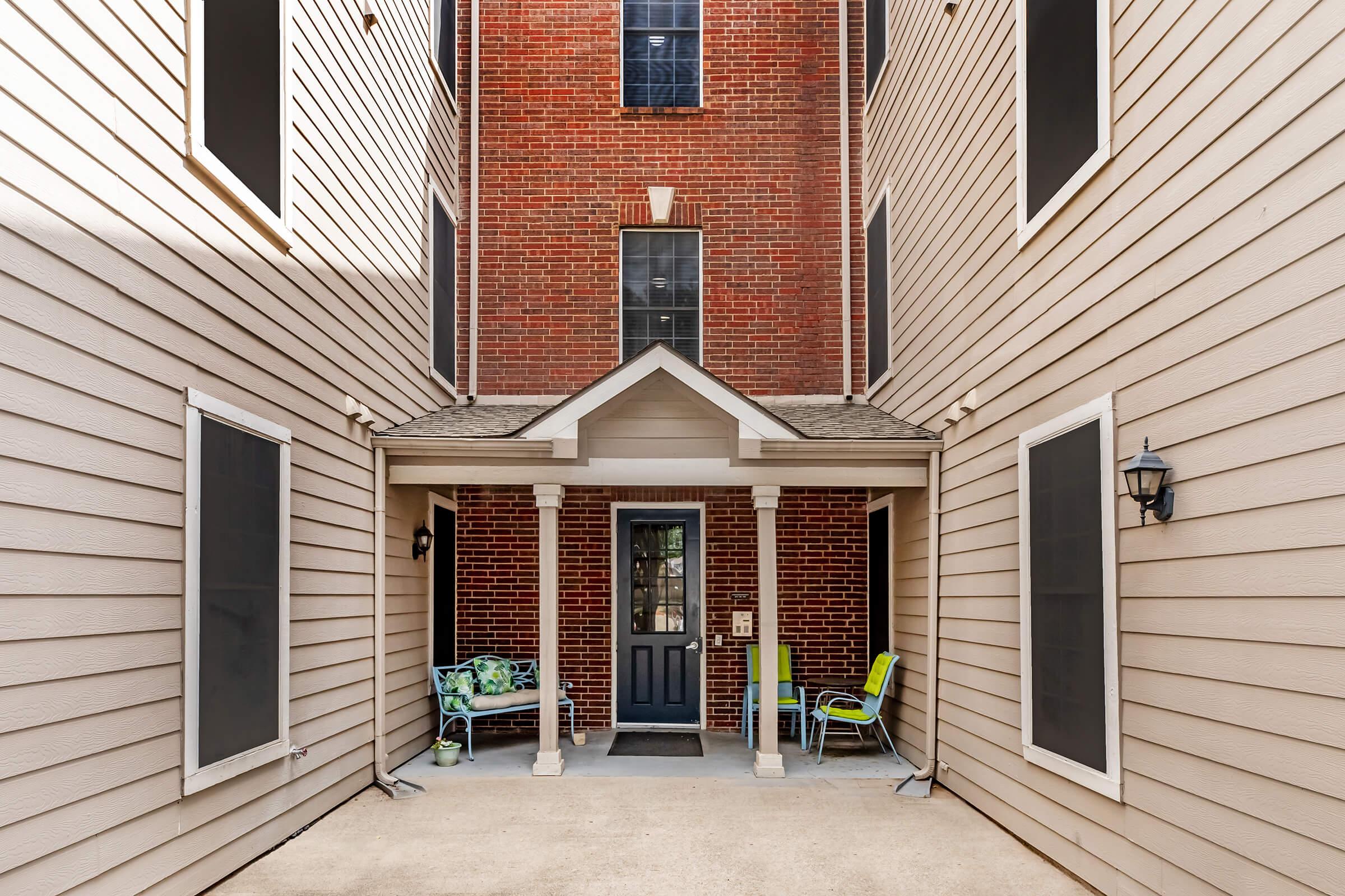 A view of a courtyard entrance to a building, featuring a front door framed by two columns. The walls are a mix of red brick and beige siding. On one side, there are two chairs and a colorful blanket, while the opposite side has a light fixture and windows with black frames. Sunlight casts shadows on the ground.
