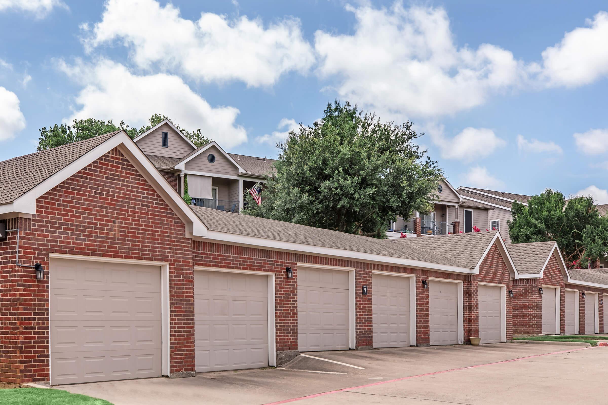 Row of brick garages with beige doors, set against a backdrop of sky and cloud. In the distance, a multi-story building is visible, partially obscured by leafy trees. The atmosphere is calm and suburban, suggesting a residential area.