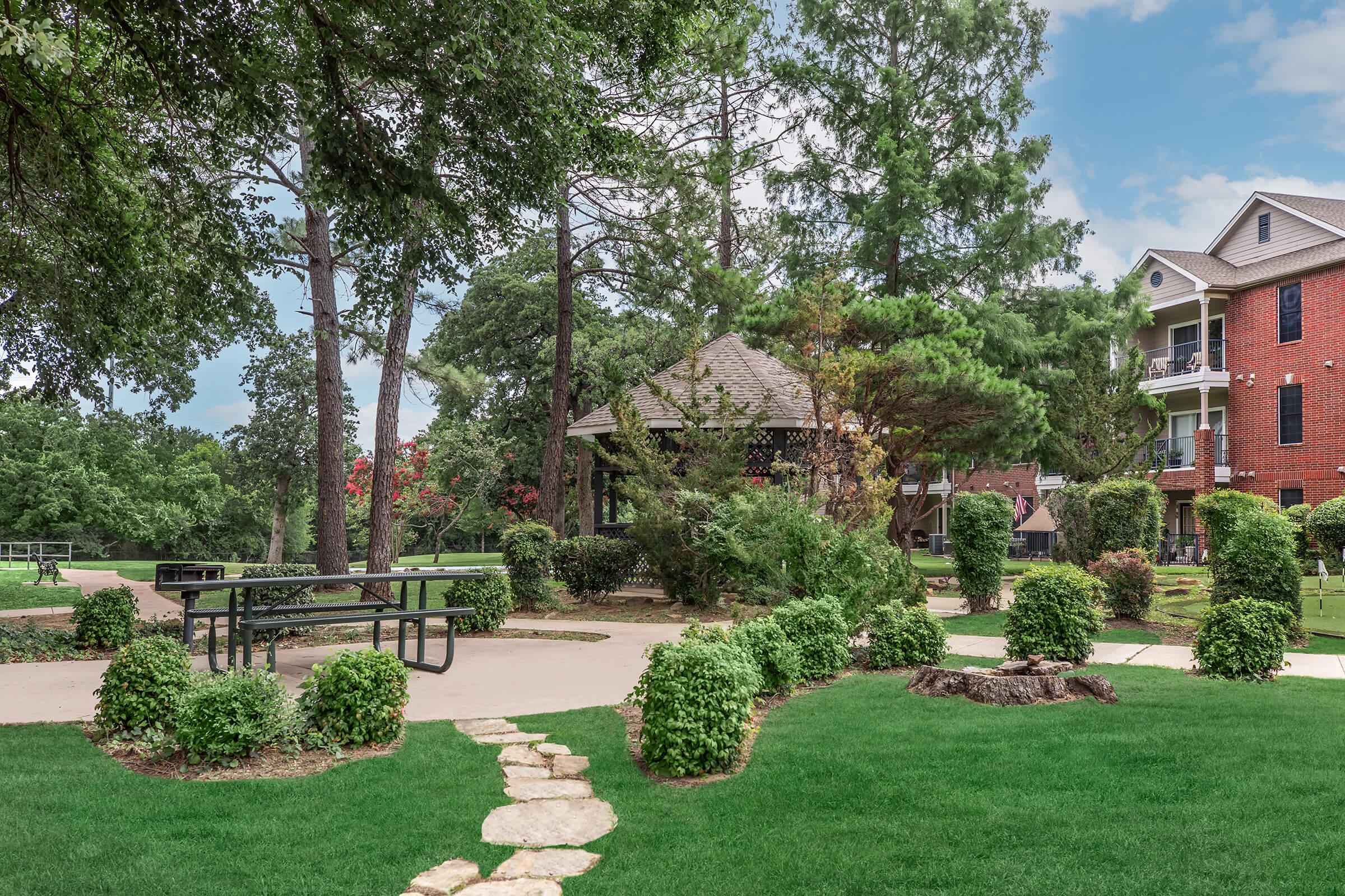 A landscaped garden featuring a gazebo surrounded by lush greenery, neatly trimmed shrubs, and a paved walkway. There is a picnic table under the gazebo, and a mix of trees in the background. A brick building is visible on the right, enhancing the serene outdoor setting.