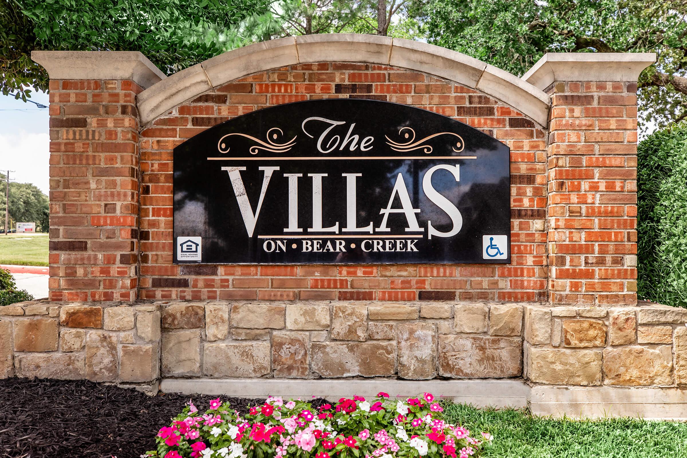 Sign for "The Villas on Bear Creek," featuring elegant typography on a black background with a brick and stone border. Floral arrangements in front add color to the scene. The sign includes a symbol indicating accessibility.