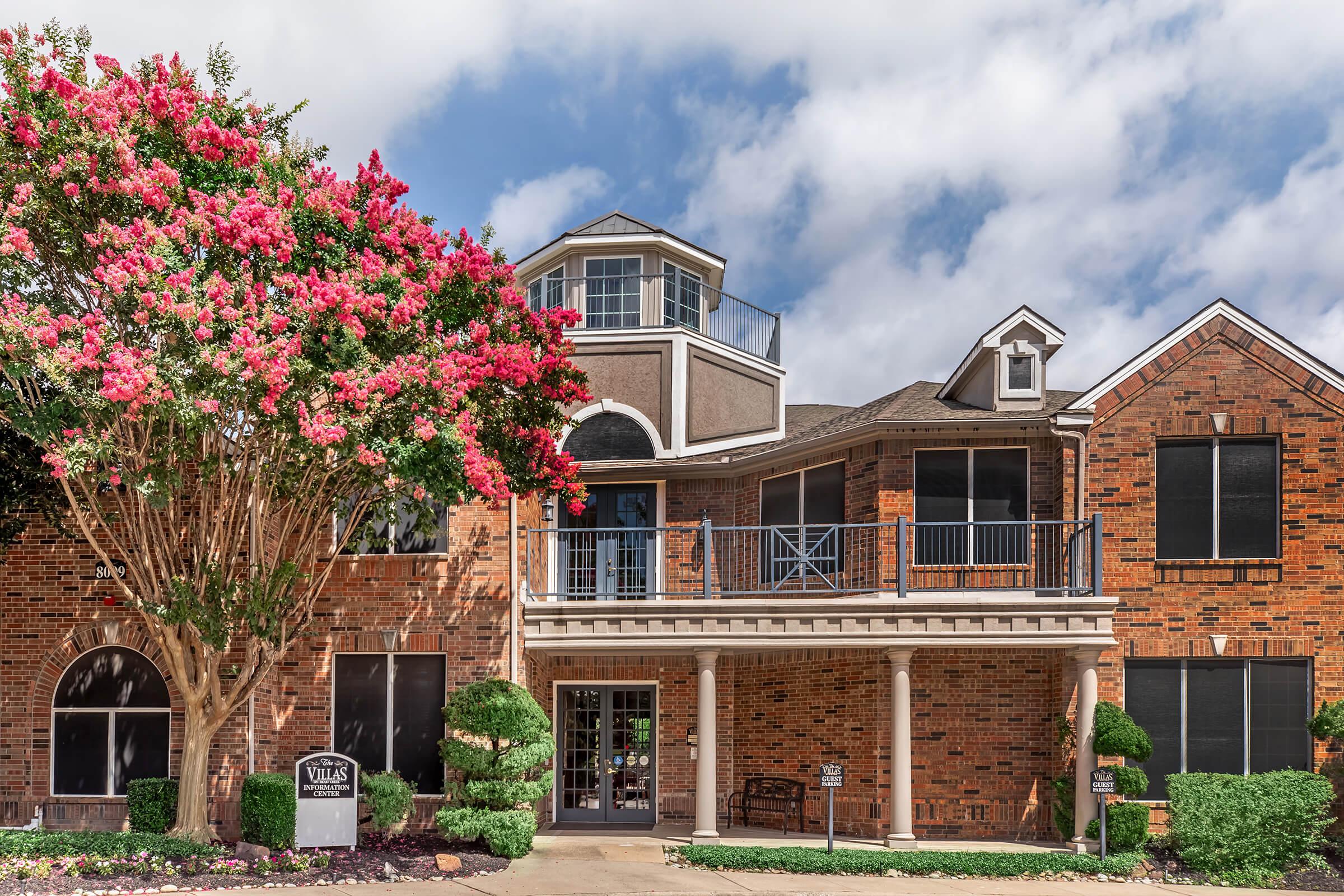 A brick building with a landscaped entrance featuring a flowering tree and shrubs. The structure has multiple levels, large windows, and a balcony. The front area includes a sign, and the sky is partly cloudy.