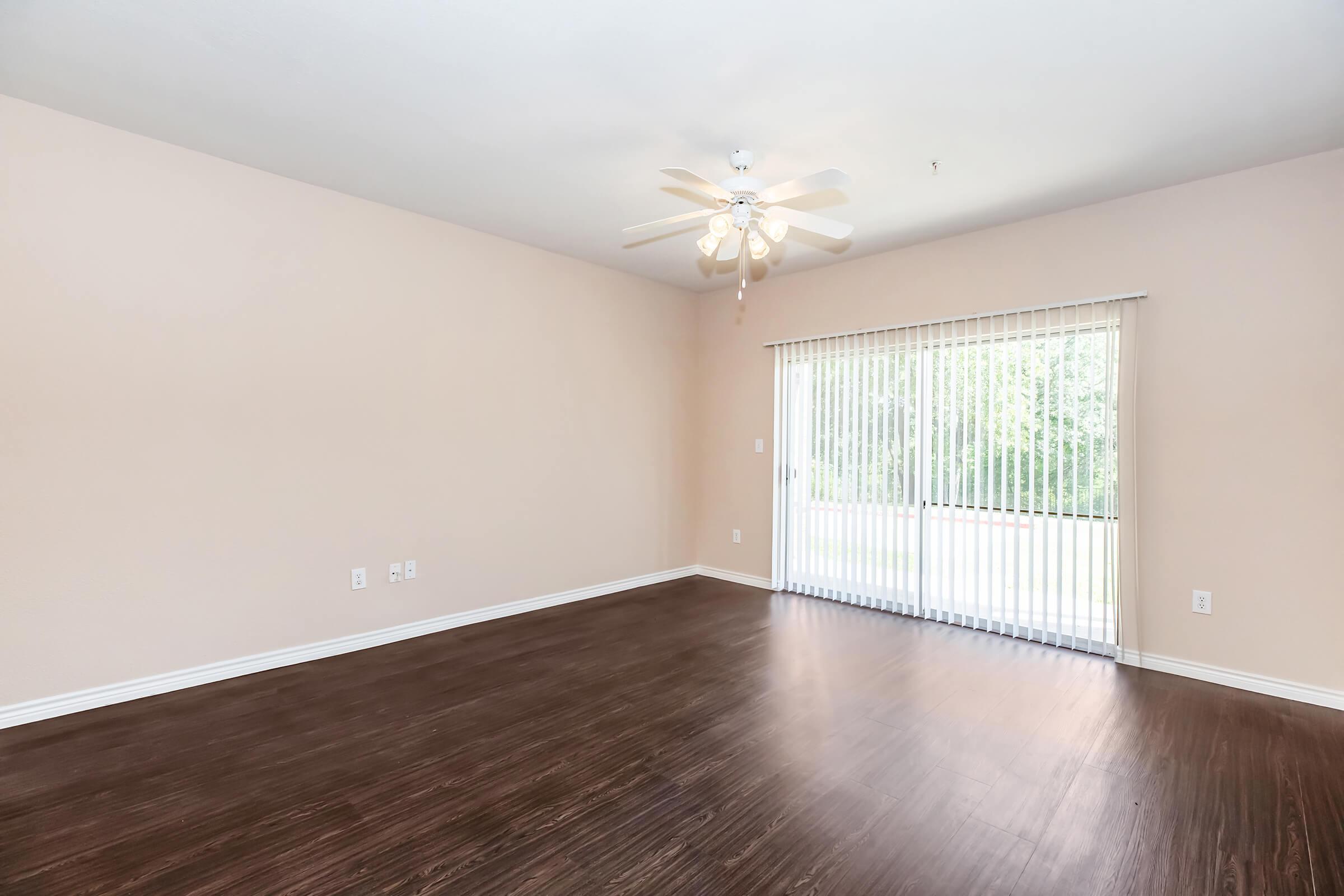 A spacious, empty room with light brown walls and dark wooden flooring. A ceiling fan is installed, and there are vertical blinds on a large window, allowing natural light to enter. The room appears bright and inviting, perfect for personal customization.