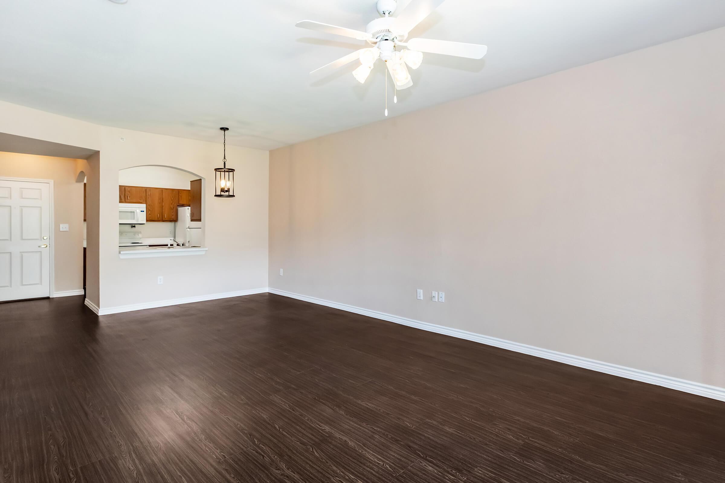 A spacious, empty living room with light-colored walls and dark wood-like flooring. A ceiling fan is visible above, and a small kitchen area is seen in the background through an archway. The room is well-lit, creating a bright and airy atmosphere.