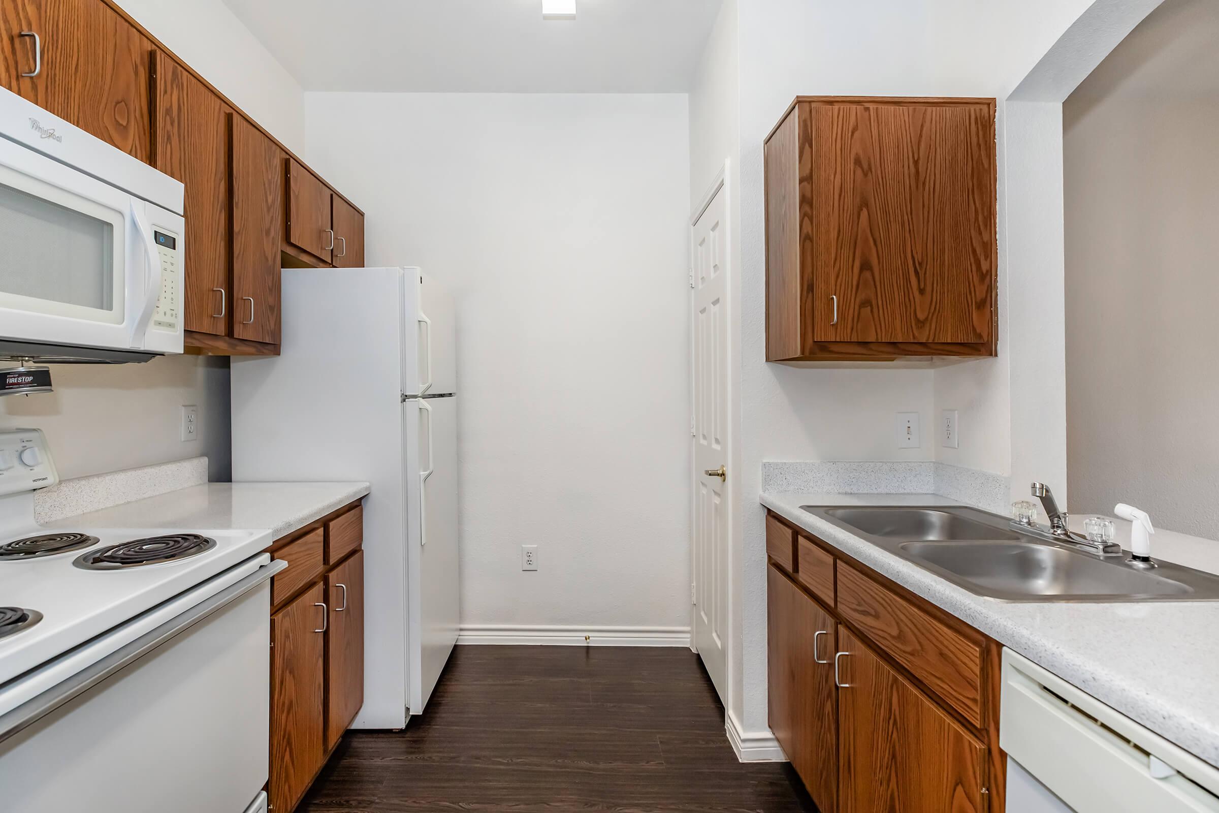 A modern kitchen featuring brown wooden cabinetry, a white refrigerator, and a white stove with an oven. The countertops are light-colored, and there is a double sink next to the stove. The walls are painted white, and the floor is a dark wood finish, creating a clean and bright space.