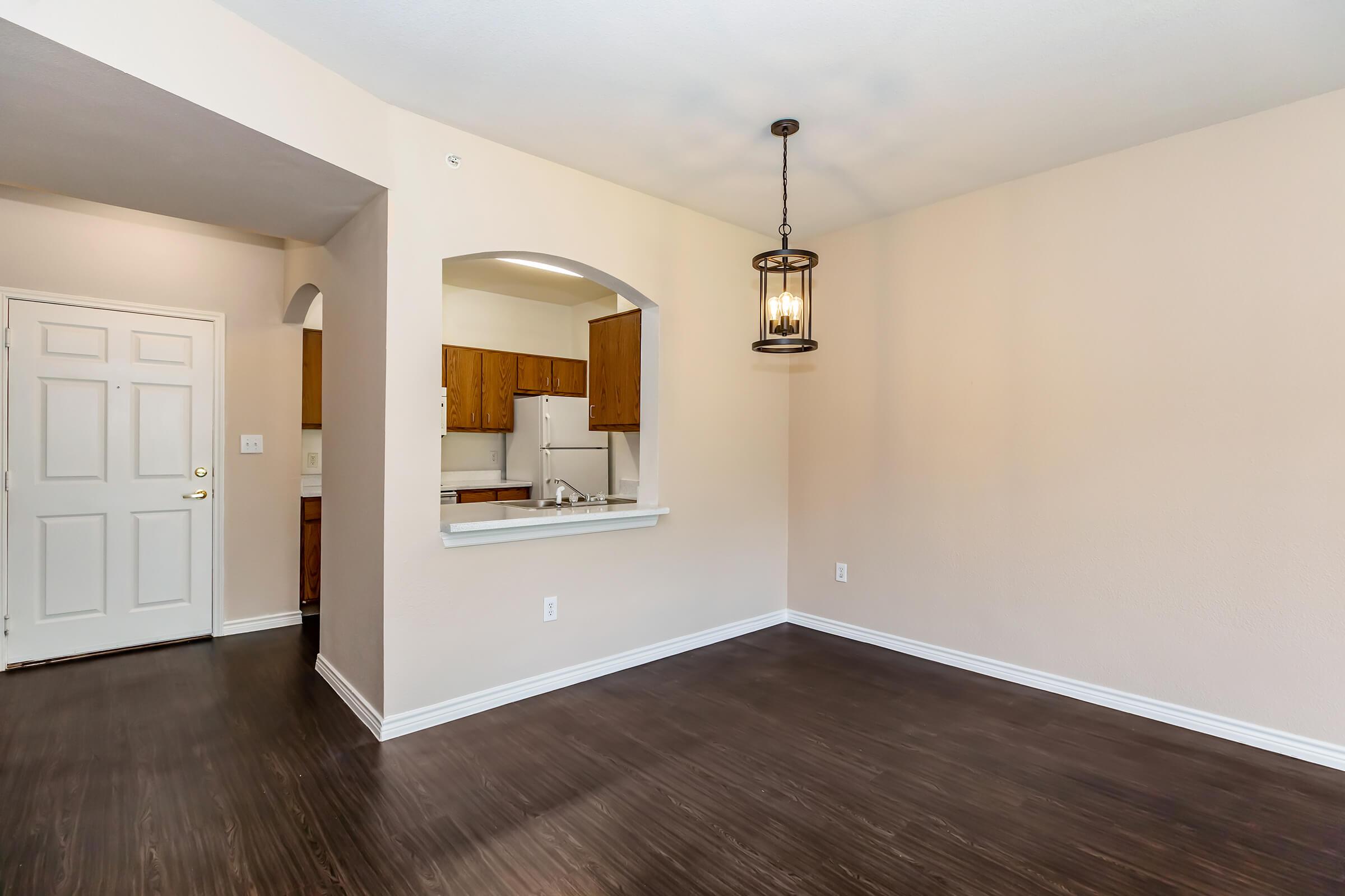 A spacious, unfurnished living room with light beige walls and dark wooden flooring. There’s a decorative light fixture hanging from the ceiling and an archway leading to a kitchen area with wooden cabinets visible in the background. A white door is on the left, leading to an entrance.