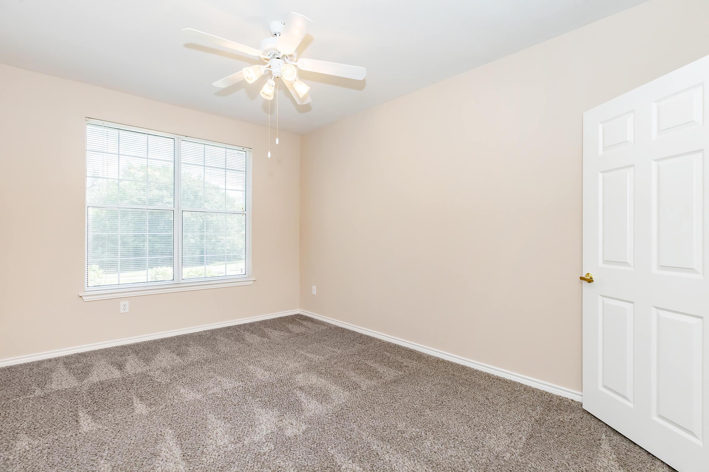A vacant room with light beige walls and carpet. It features a ceiling fan with five blades and a large window with white blinds, allowing natural light to enter. A closed white door is on the right, and the space appears clean and ready for decoration or furniture.