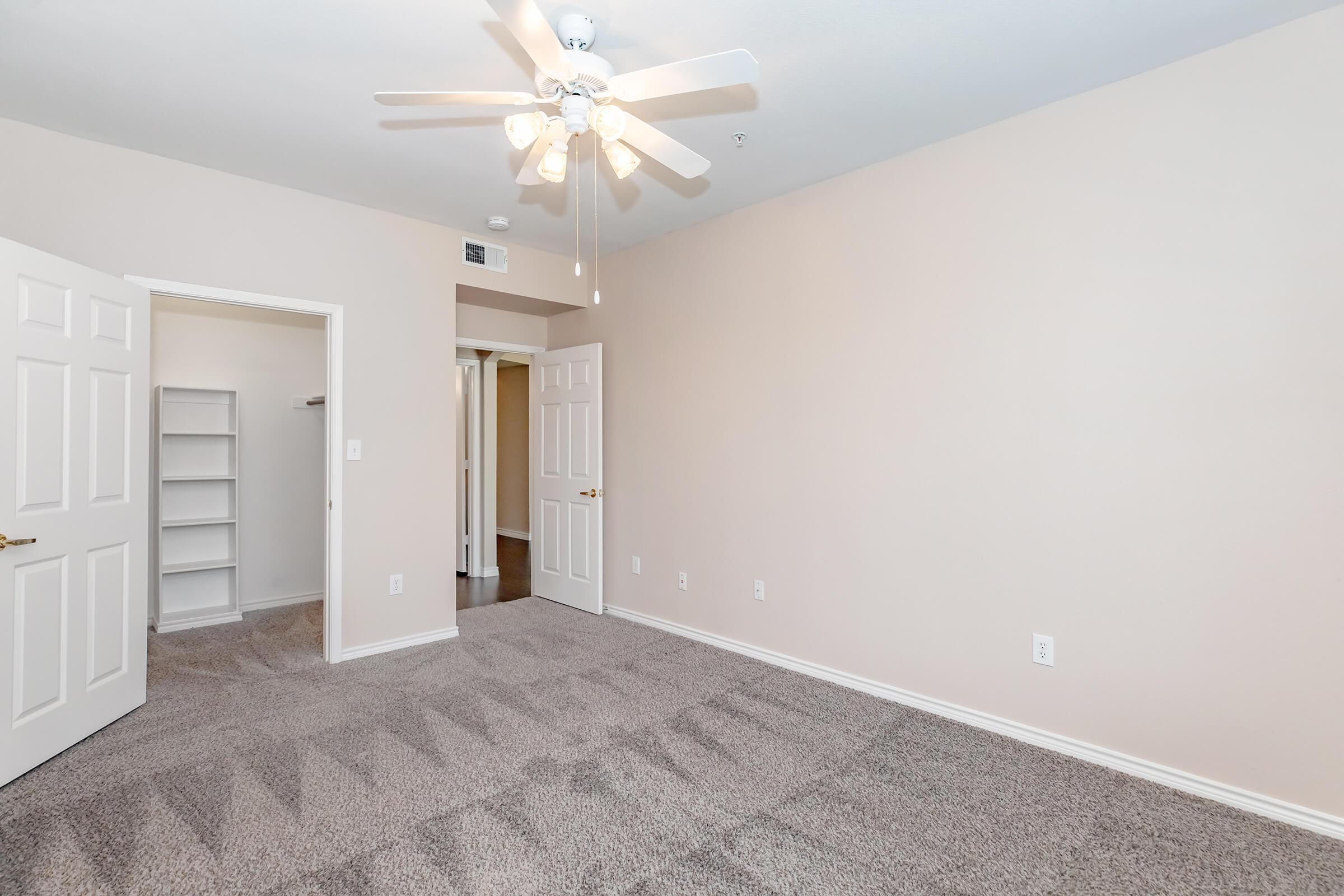 A light-filled room with beige walls and plush carpeting. It features a ceiling fan with multiple blades, two open doors leading to adjacent areas, and a closet with built-in shelves visible. The space has a clean, neutral aesthetic, suitable for a bedroom or living area.