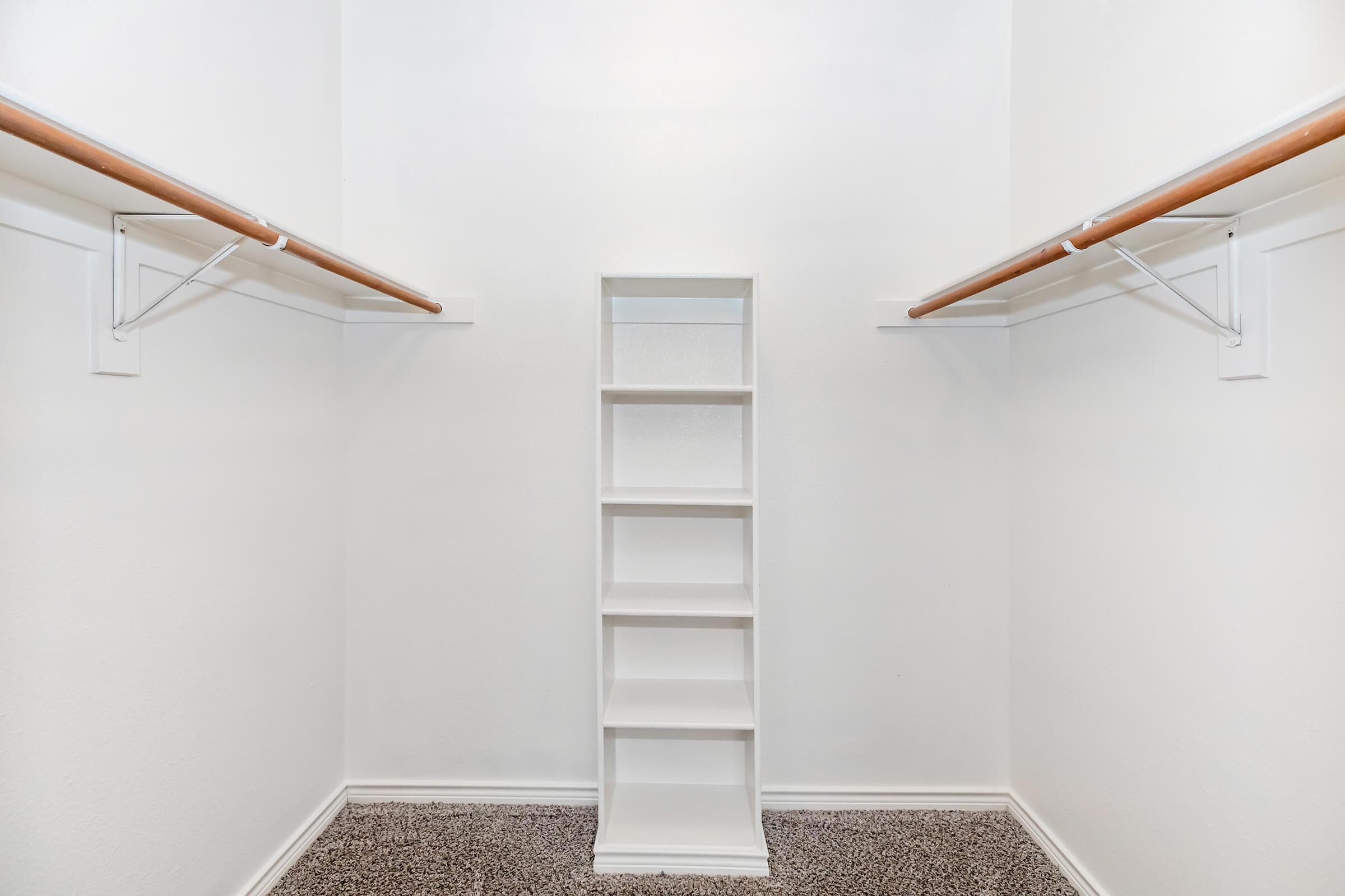 A spacious empty closet featuring two hanging rods on either side and a central set of white shelves. The walls are painted white, and the floor is covered with gray carpeting, creating a clean and organized appearance.