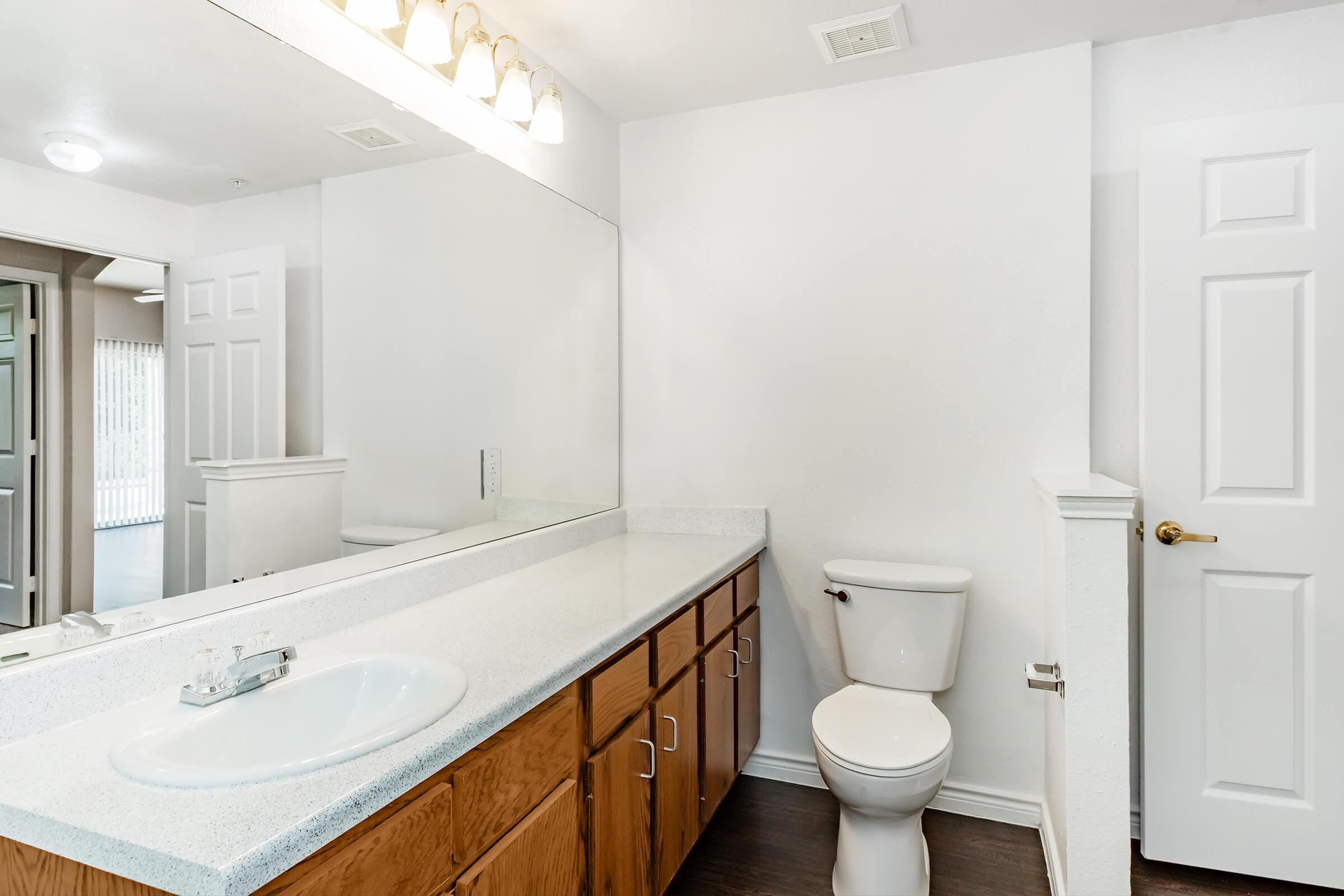 A bright and clean bathroom featuring a large mirror above a sink with a countertop, wooden cabinets below, a white toilet, and a door leading to another room. Soft lighting from a fixture above illuminates the space, which has white walls and dark flooring.
