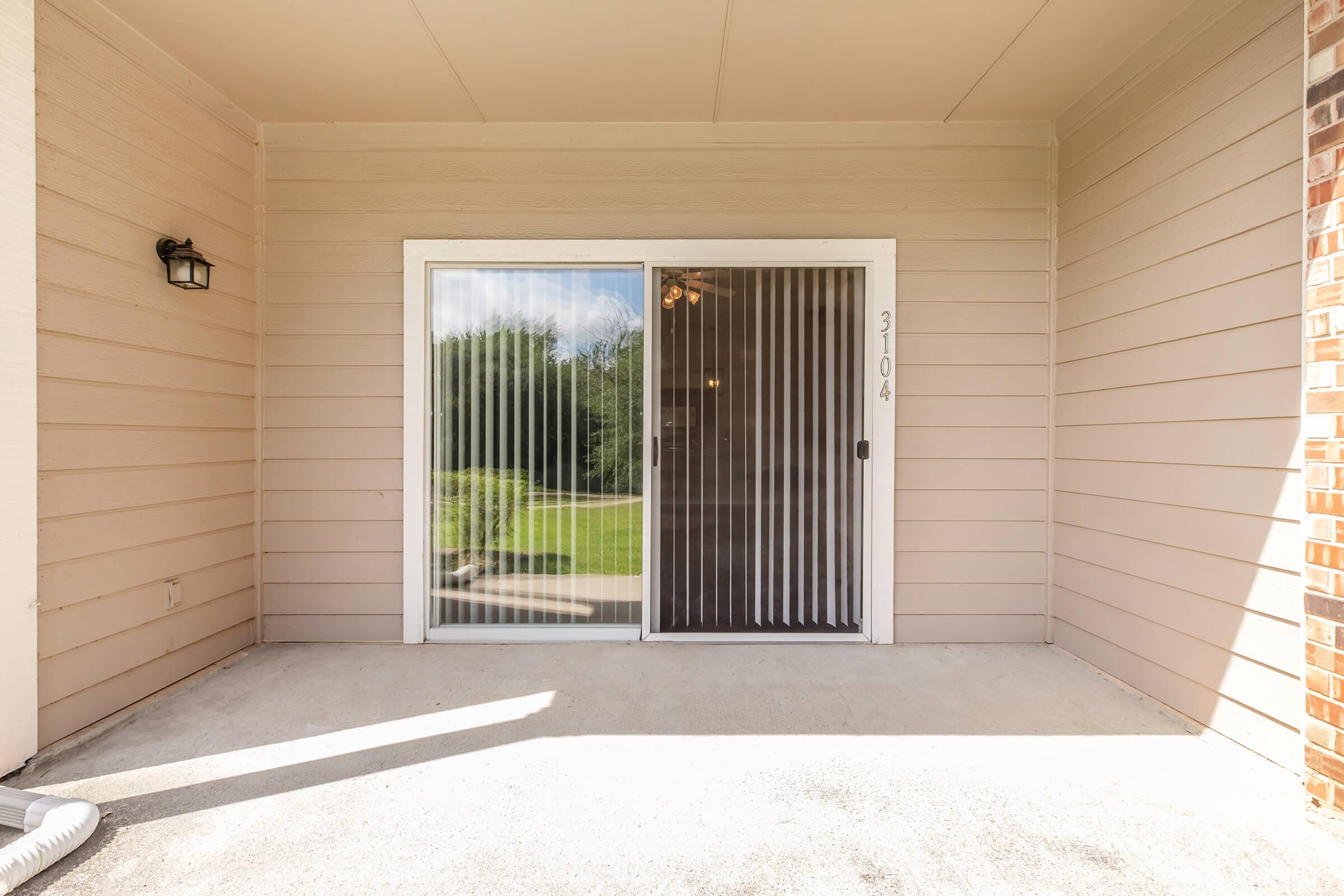 A clear sliding glass door with vertical blinds, framed by beige siding on either side. The door is labeled with the number "3104." The porch area is concrete with a shadow cast by the door frame, and a light fixture is mounted to the left of the door. A green lawn is visible through the door.