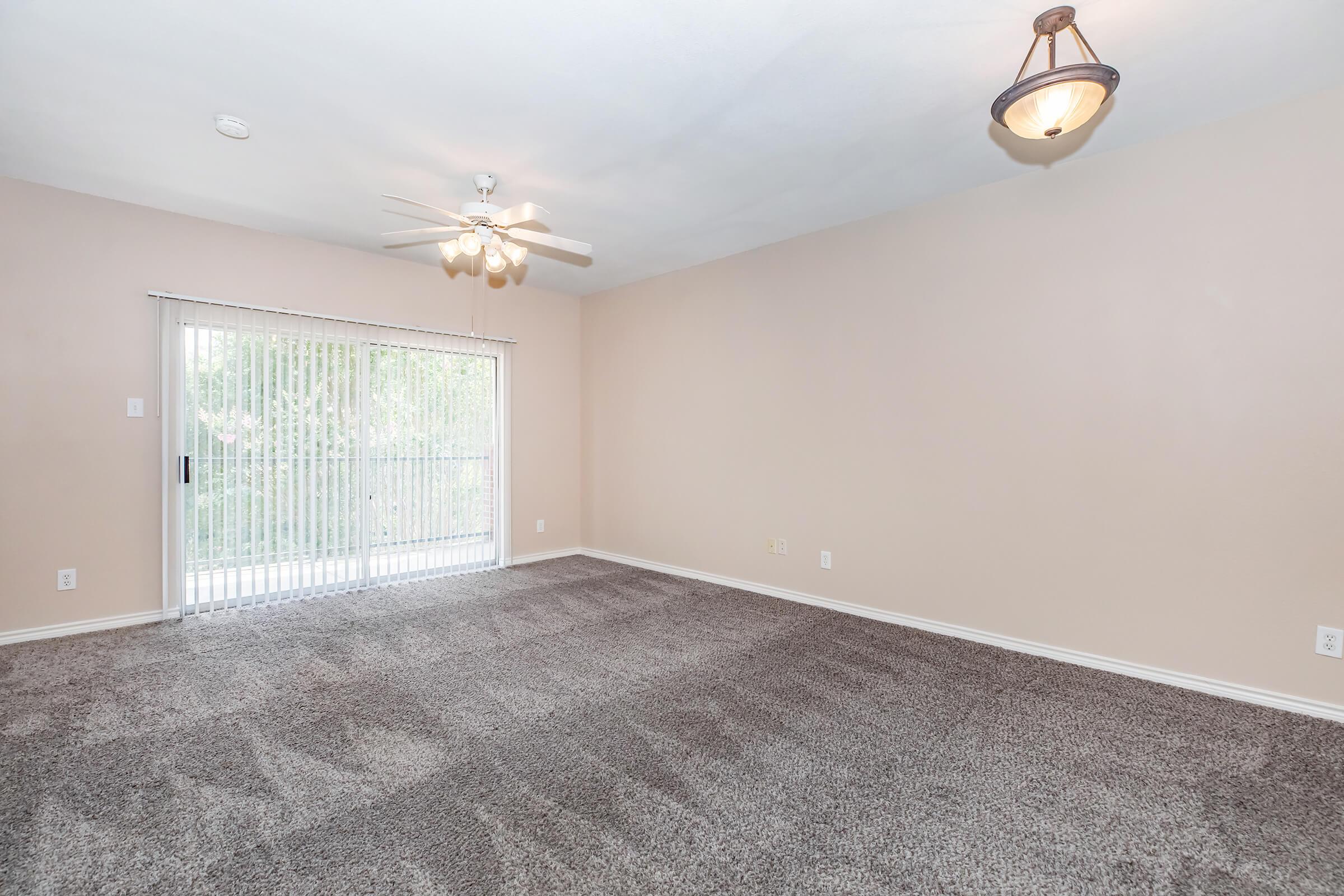 Spacious interior of a light-colored room featuring plush carpet, a ceiling fan, and a large sliding glass door leading to a balcony with vertical blinds. Soft lighting enhances the inviting atmosphere, making it suitable for various uses such as a living area or bedroom.