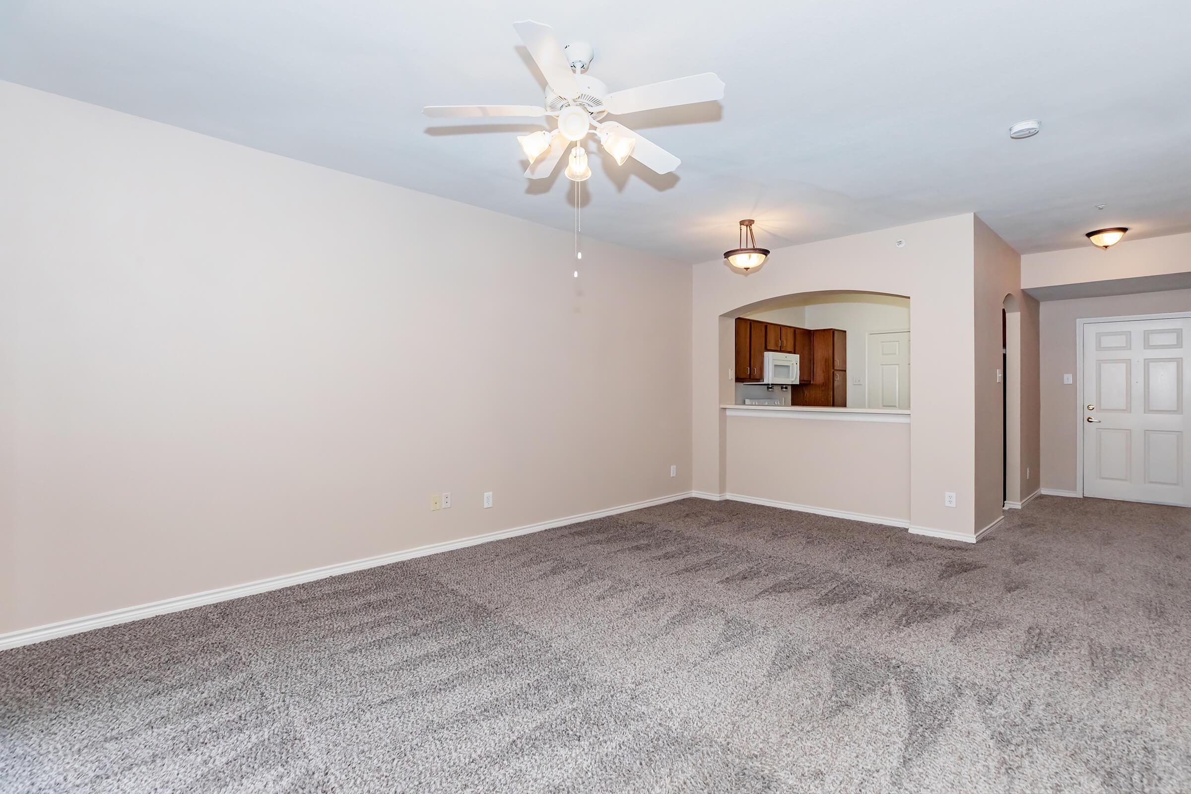 Spacious living area with light beige walls and soft carpet flooring. A ceiling fan is installed in the center, and to the left, there's an open kitchen area visible through an archway. The kitchen features light wooden cabinetry and modern appliances, complemented by warm lighting from hanging fixtures.