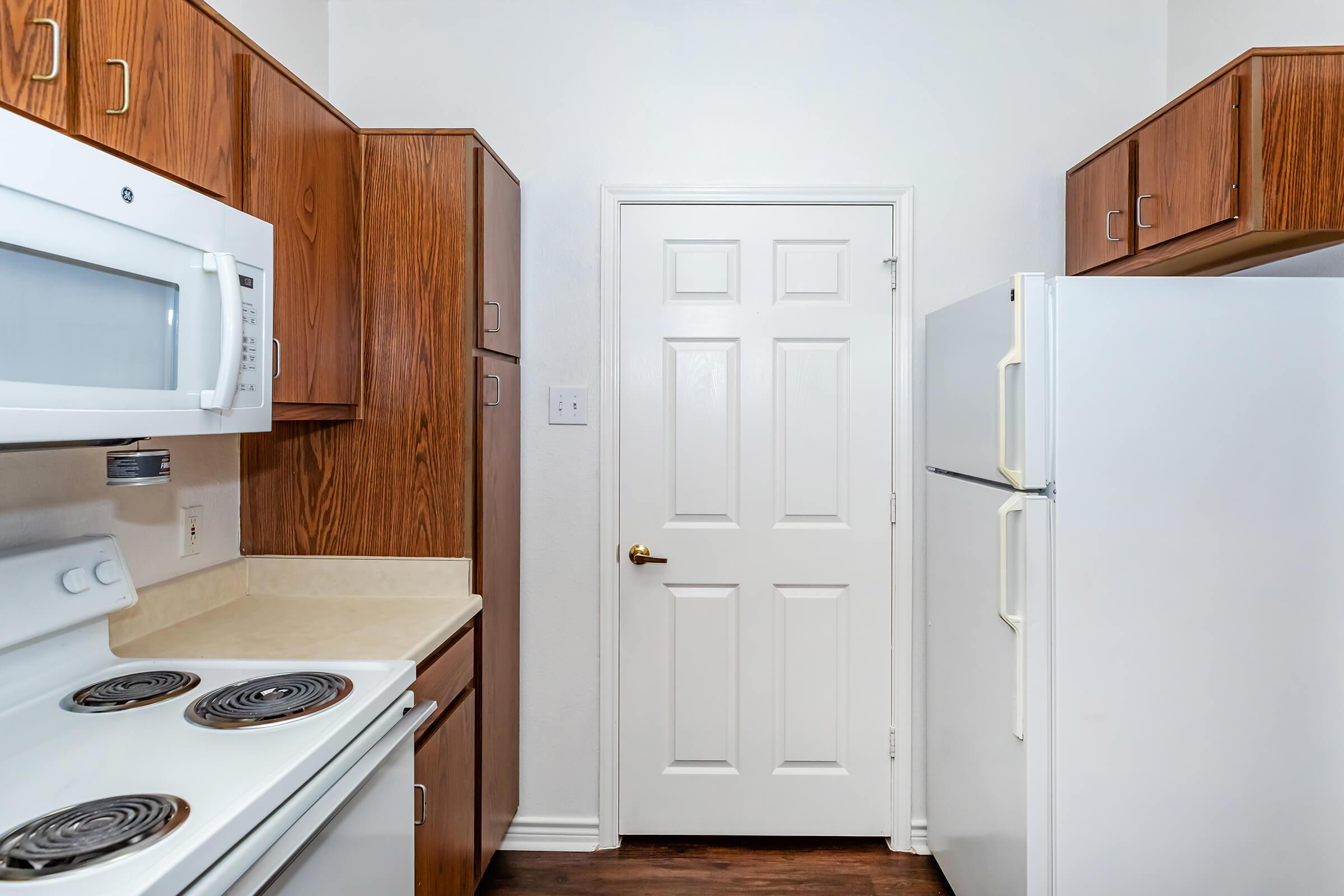 A view of a modern kitchen featuring wooden cabinets, a white stove with a built-in microwave, and two refrigerators. The room has light-colored walls and a closed white door leading to another space. The floor is dark wood, enhancing the kitchen's overall aesthetic.