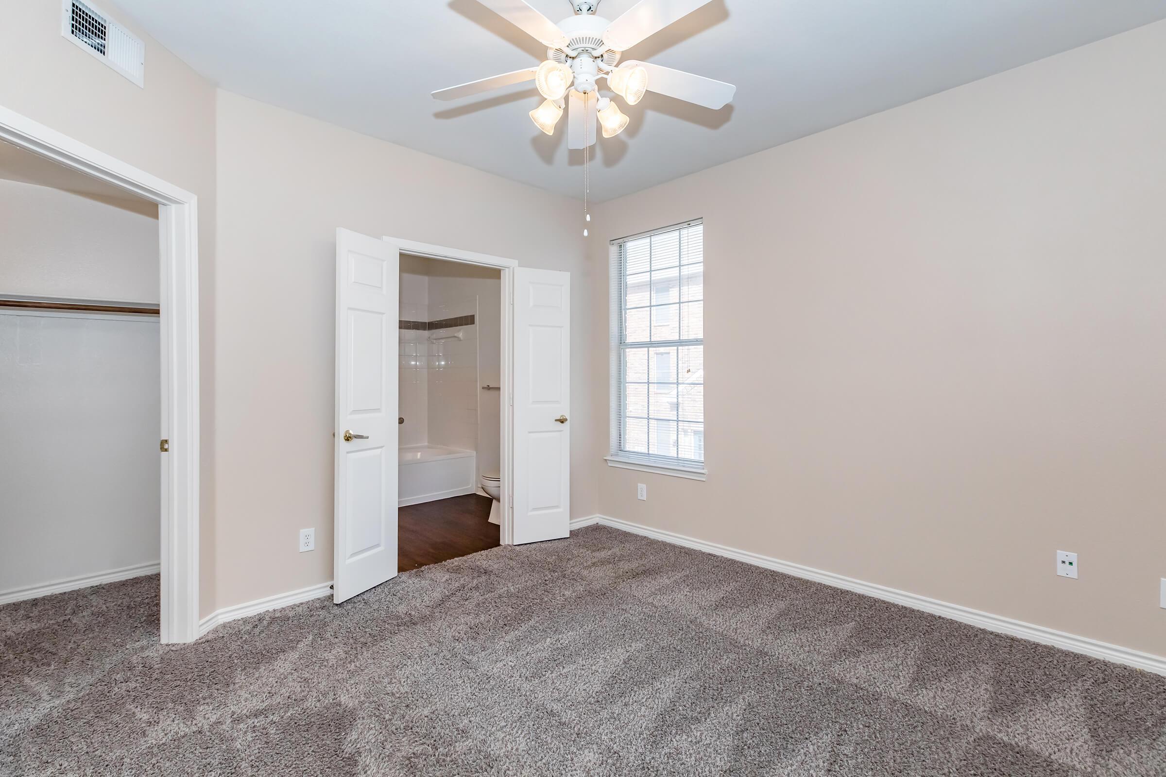 A well-lit bedroom featuring beige walls, a ceiling fan with light fixtures, and a carpeted floor. There are two doors leading to a closet and a bathroom, with a window letting in natural light. The room has a cozy, spacious feel and is freshly painted, making it inviting and ready for occupancy.