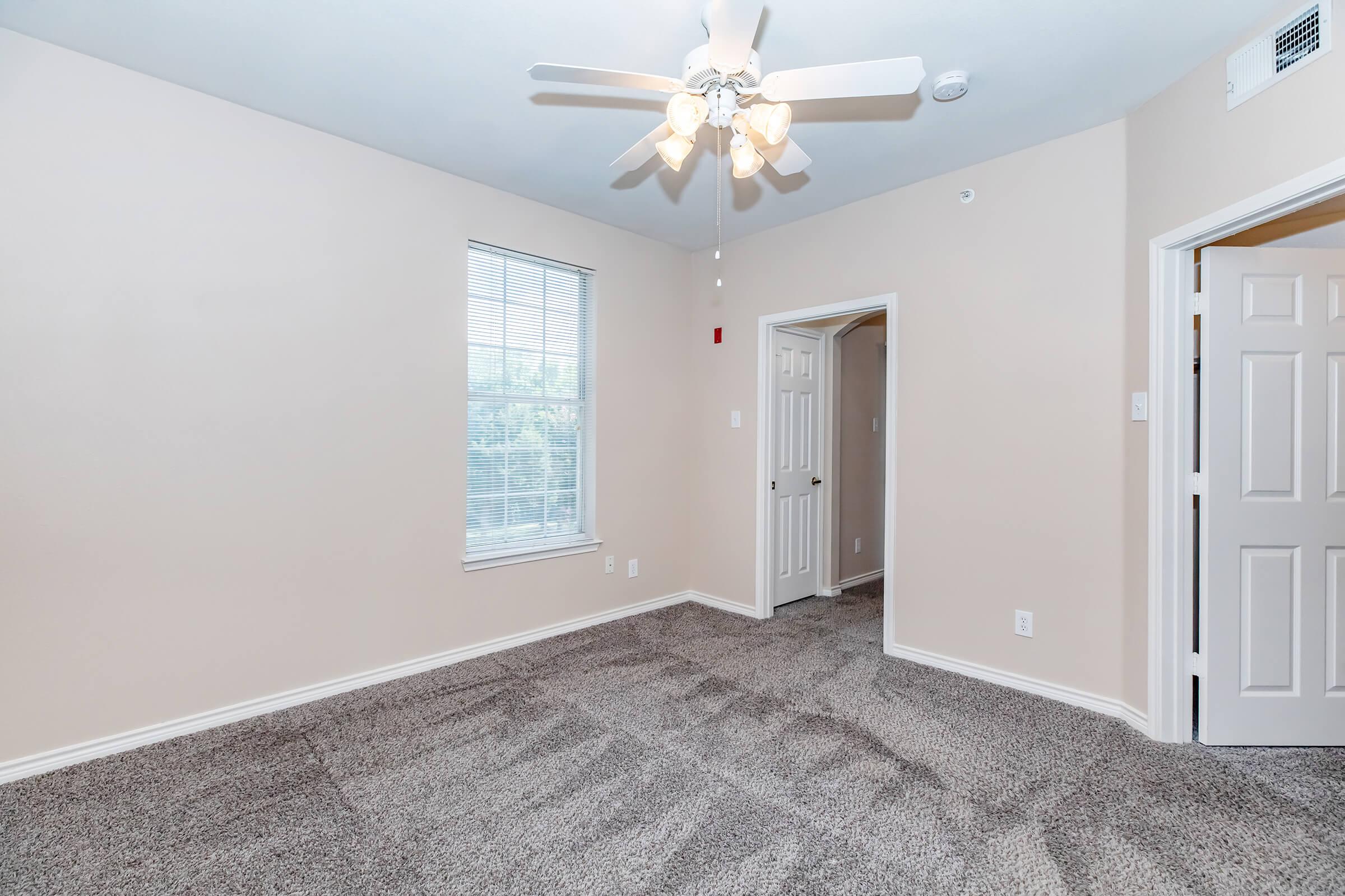 A light and airy bedroom featuring cream-colored walls and plush carpet. A ceiling fan hangs in the center, and there are two doors leading to adjacent rooms. A window allows natural light to filter in, enhancing the spacious feel of the room.