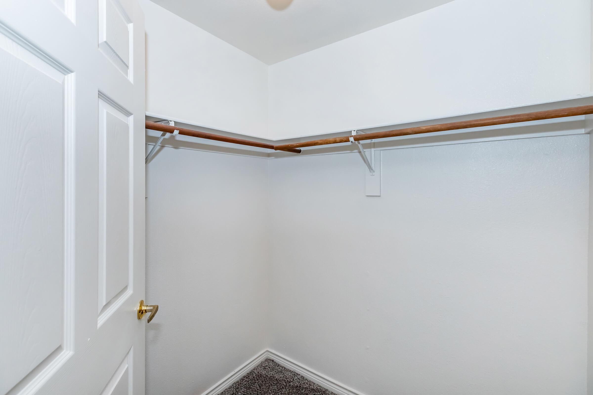 Empty closet with white walls, a single wooden hanging rod, and a carpeted floor. The door is slightly open, revealing the uncluttered space inside.