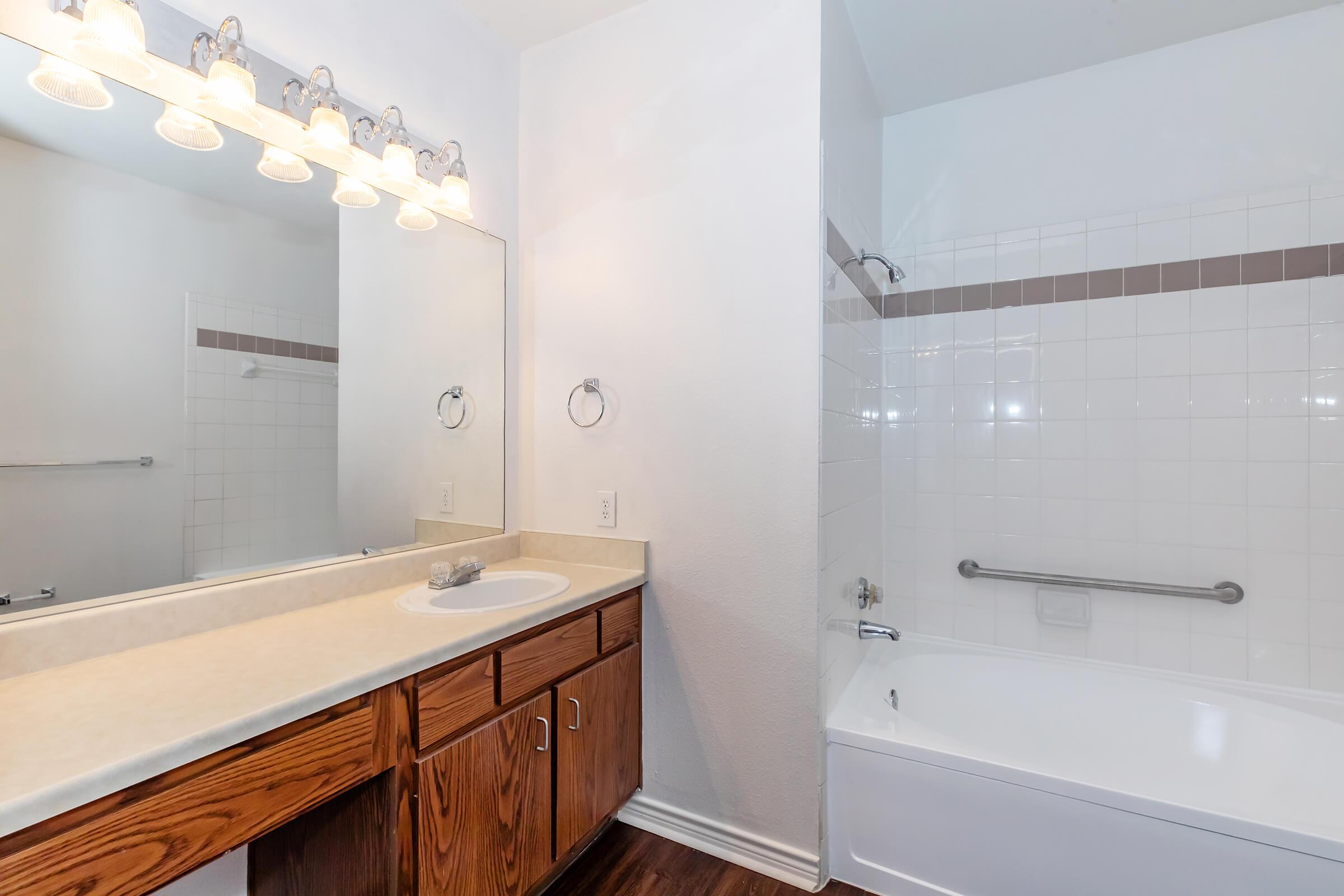 A clean, well-lit bathroom featuring a large mirror above a beige countertop with a sink, wooden cabinetry, and a tub-shower combination with white tiles. The walls are painted white, and there are towel bars and light fixtures above the mirror. The floor is dark wood.