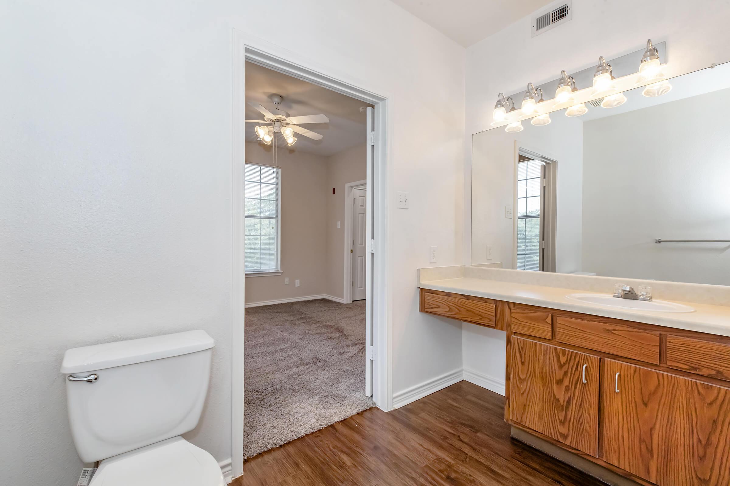 A bright bathroom featuring a white toilet and a wooden vanity with a sink, illuminated by overhead lights. A door leads to a carpeted room with a ceiling fan and large windows, allowing natural light to enter. The walls are painted in a soft, neutral color, creating a spacious and inviting atmosphere.