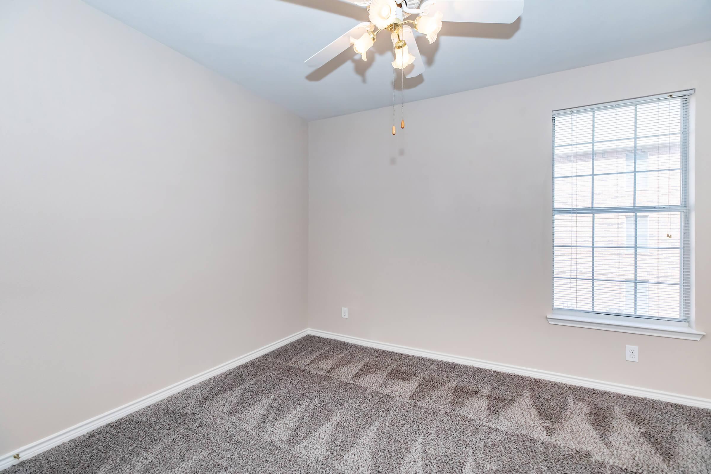 Empty room with light beige walls and a light fixture on the ceiling. The floor is covered with gray carpet, and there is a window with white blinds letting in natural light.