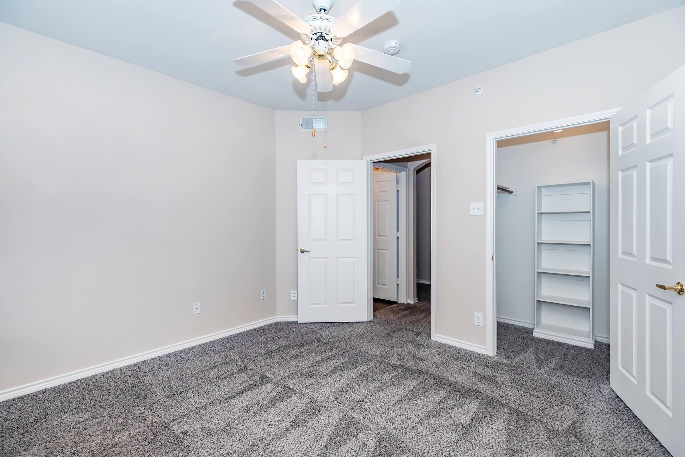 A well-lit bedroom featuring light beige walls, a ceiling fan with five bulbs, and soft gray carpet. The room has two white doors leading to a closet and another space, with a built-in shelf visible in the closet area. The overall atmosphere is spacious and inviting.