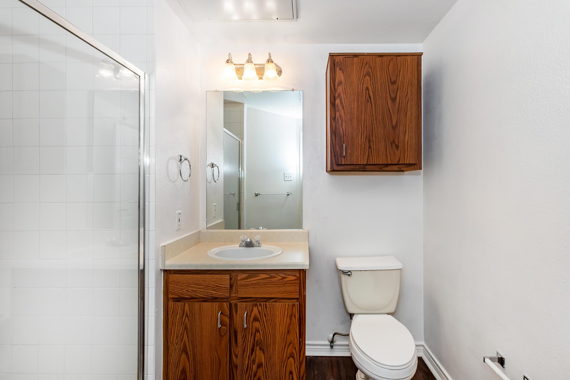 A clean, modern bathroom featuring a shower area with glass doors, a single sink with wooden cabinet storage, a mirror above the sink, and a toilet. The walls are painted white, and the flooring is a dark wood laminate. There is ample natural light coming from an adjacent room.