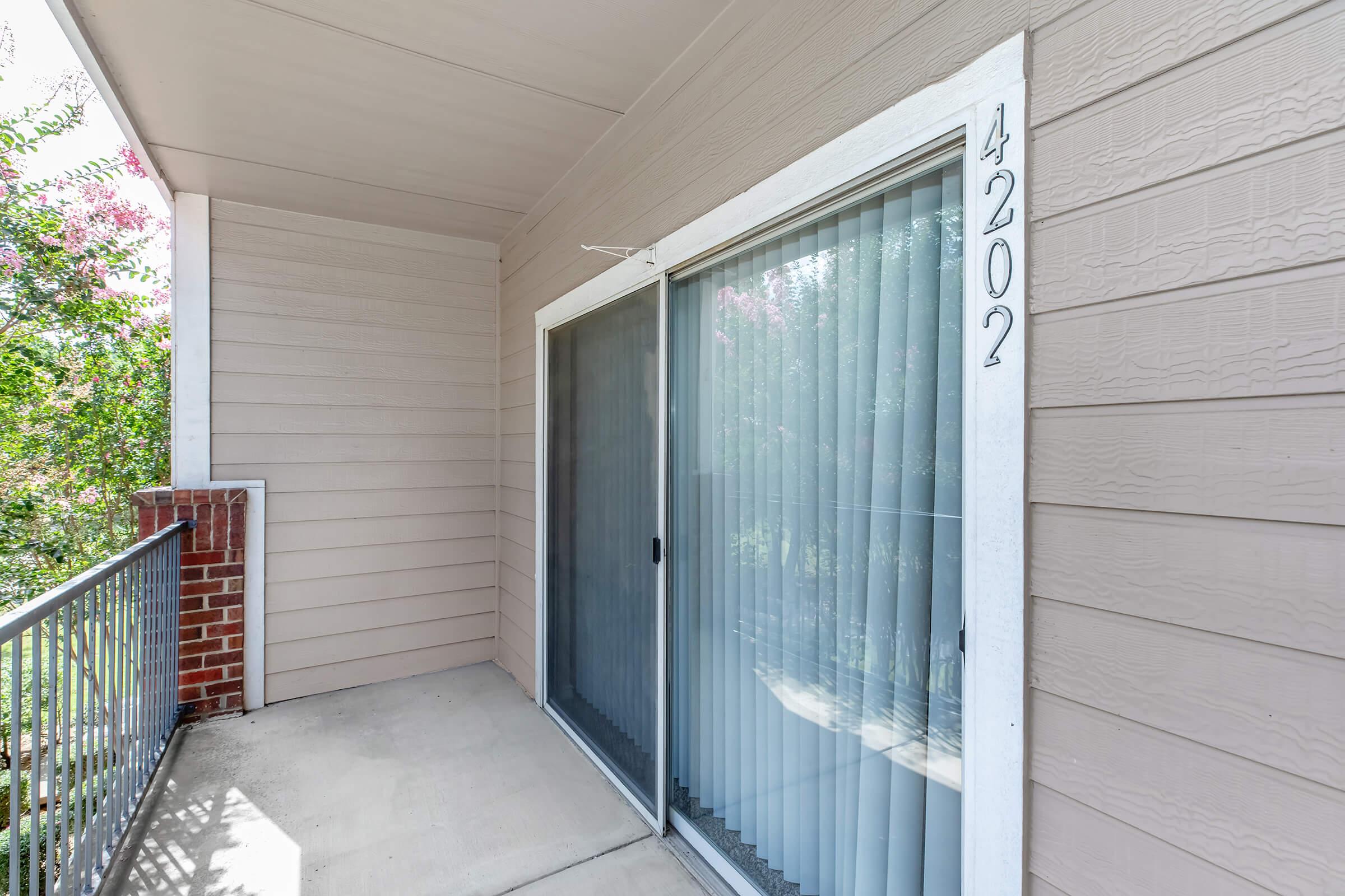 View of a residential balcony area featuring sliding glass doors with sheer curtains, a concrete floor, and light-colored siding. The number "4202" is displayed on the wall next to the doors. Surrounding greenery is visible through the transparent curtains, adding a touch of nature to the space.