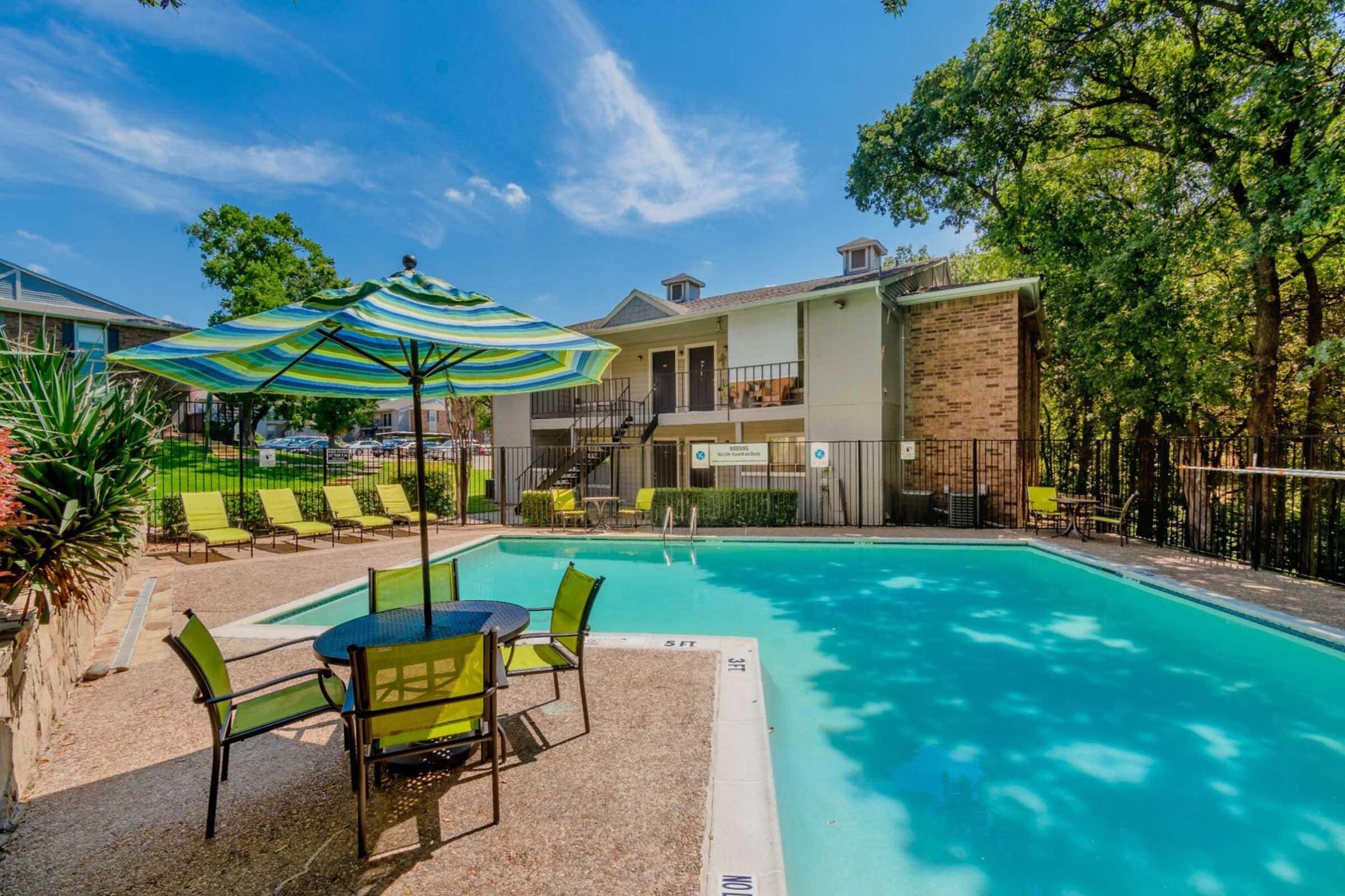 A bright outdoor pool area featuring a clear blue swimming pool surrounded by a concrete deck. There are several green lounge chairs and a table with umbrellas for shade. In the background, there is a multi-story building and lush trees providing a natural setting.