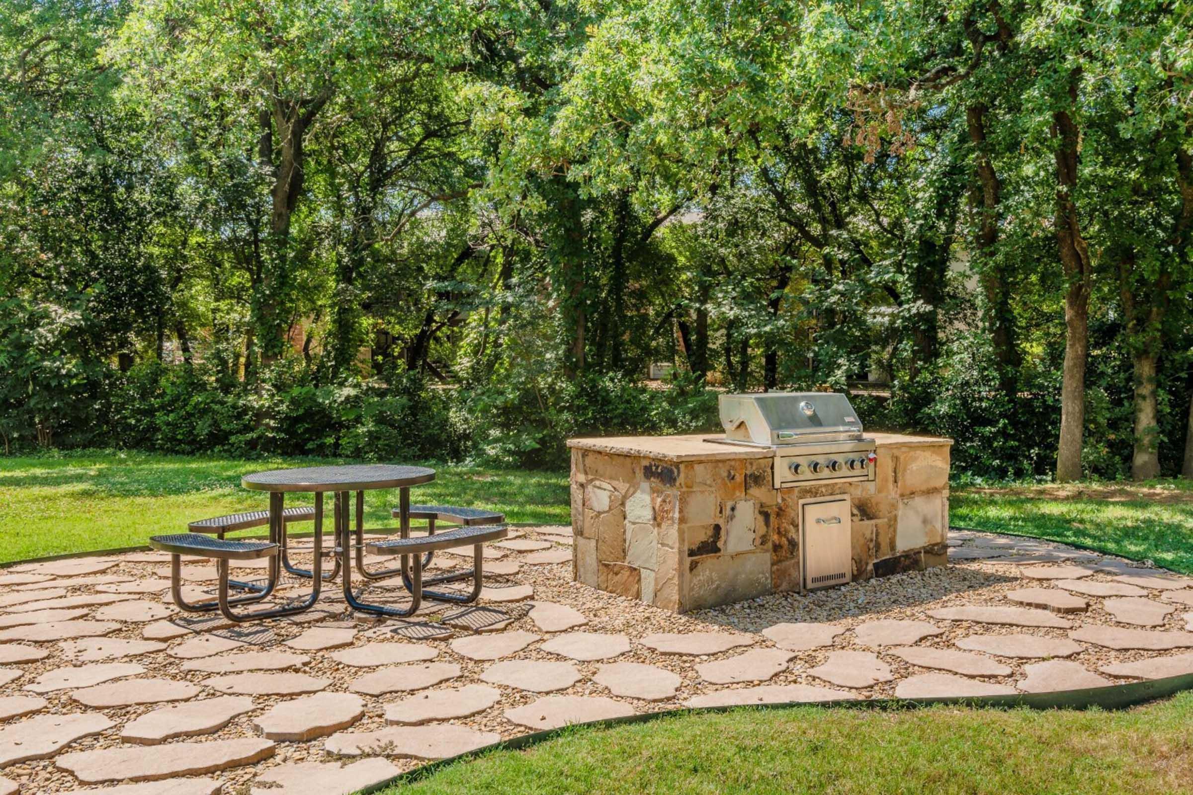 Outdoor barbecue area featuring a stone grilling station and a circular picnic table surrounded by greenery. The setting includes a textured stone pathway and trees in the background, creating a relaxing atmosphere for outdoor cooking and dining.