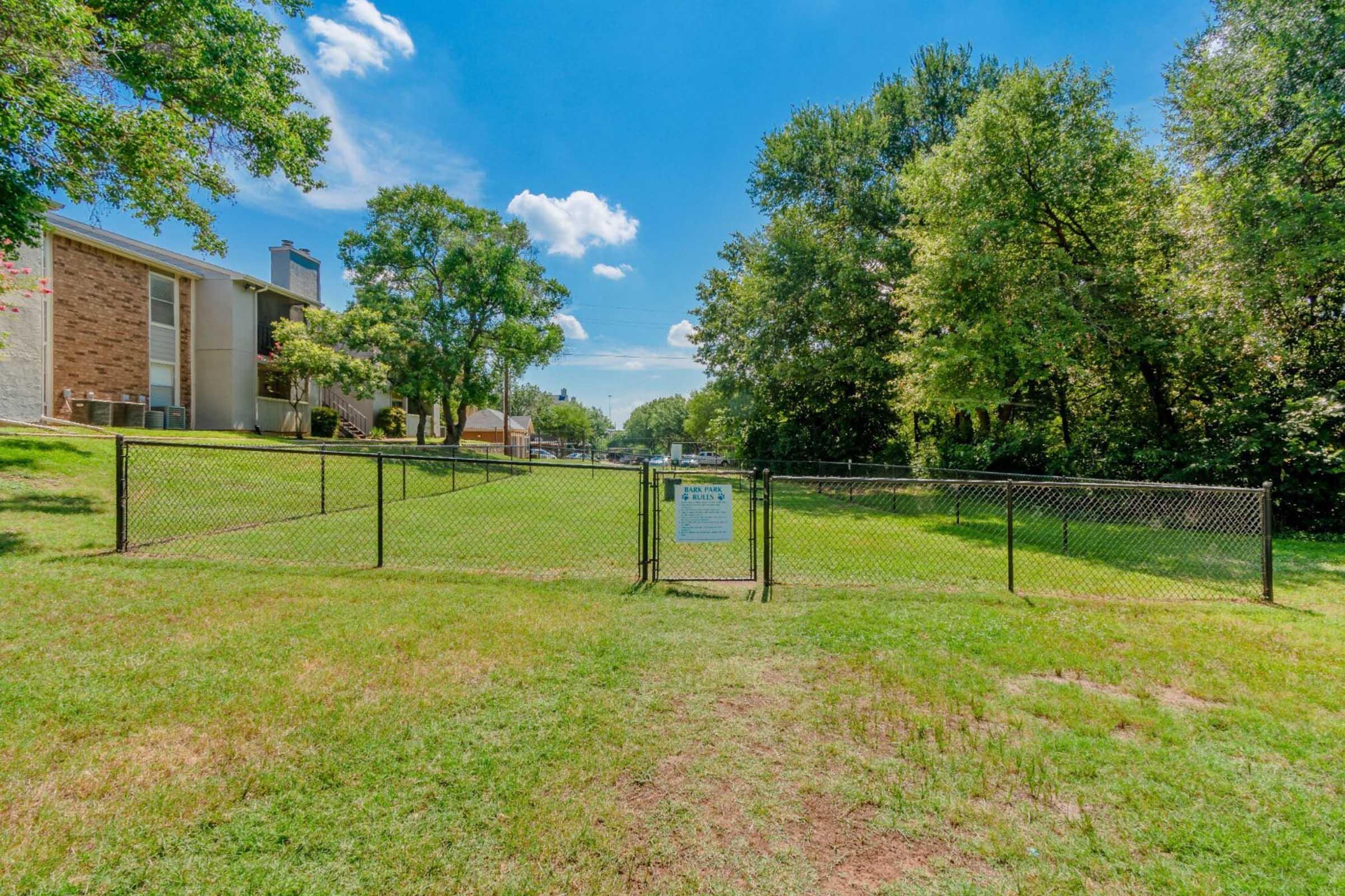 A fenced grassy area with a sign visible in the center. The background features a few trees and an apartment building. The sky is clear with a few clouds, and the area is well-lit, suggesting a sunny day.
