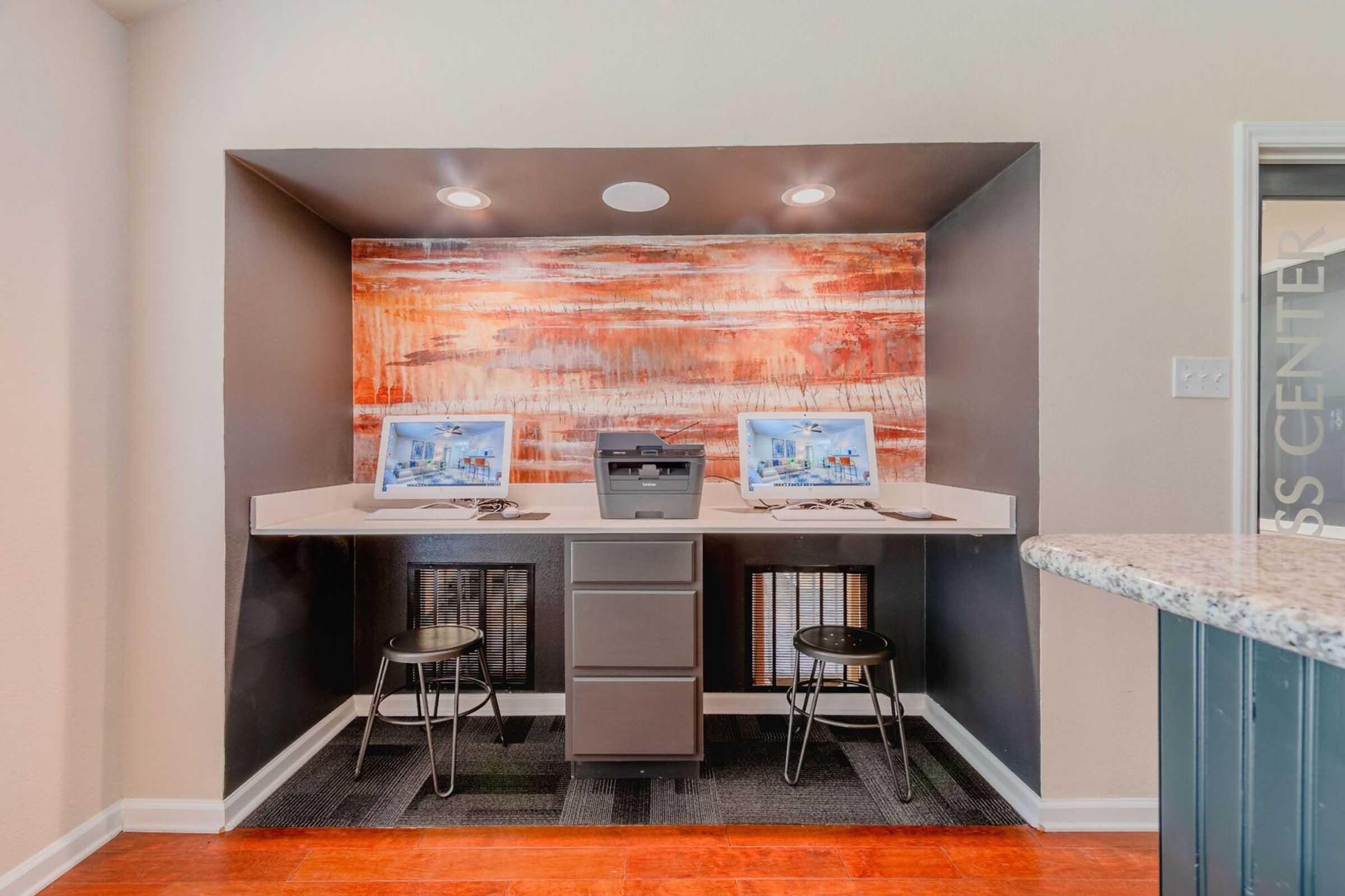 A modern business center featuring two desktop computers on a shared workspace, a printer, and ergonomic stools. The backdrop has an abstract orange and black design. The setting is well-lit and designed for productivity, with a granite counter and a stylish interior.