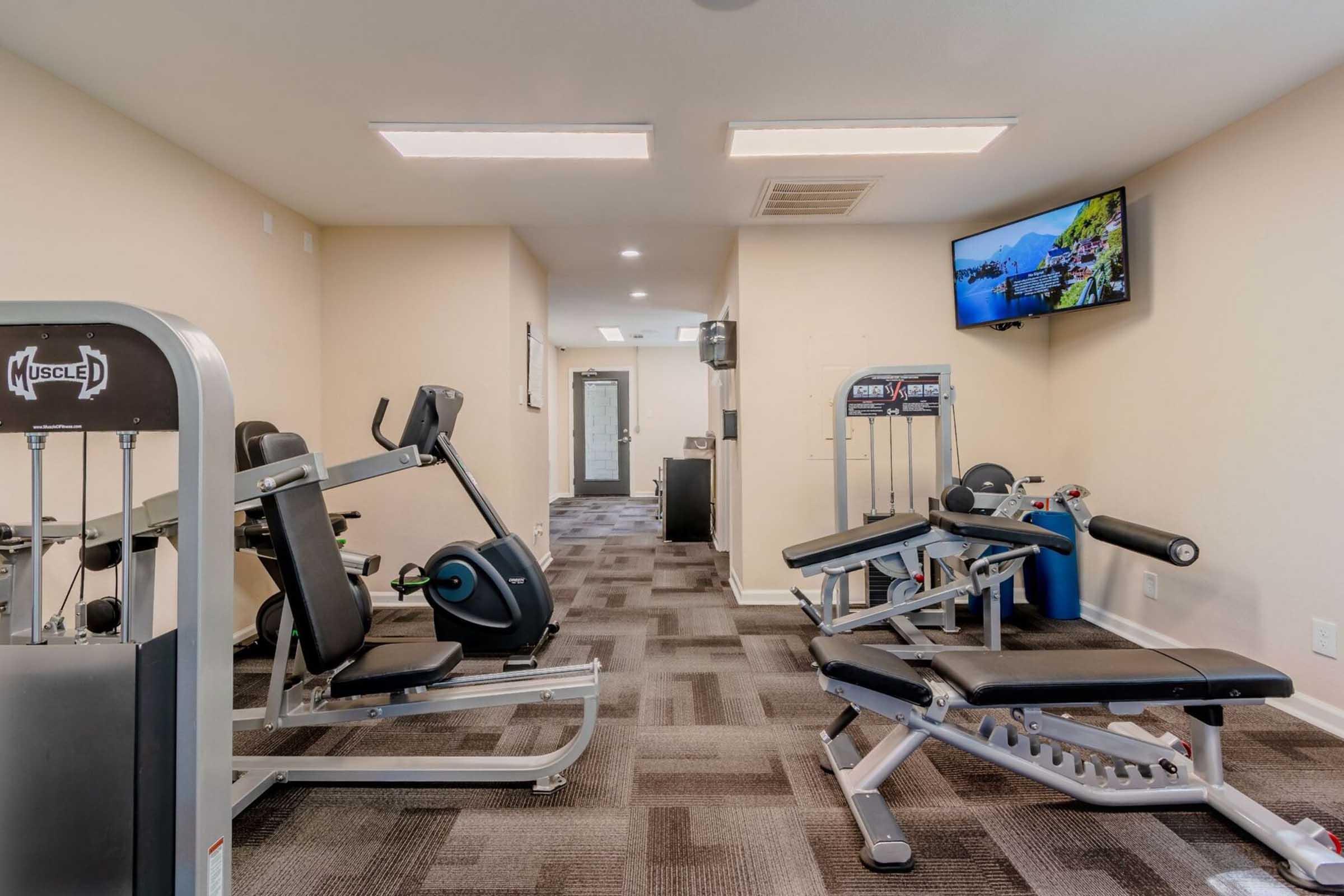 A small gym area featuring various exercise equipment, including a stationary bike, weight machines, and a TV mounted on the wall. The space is well-lit with neutral-colored walls and a clean, modern appearance. Fitness equipment is neatly arranged on a patterned carpet.