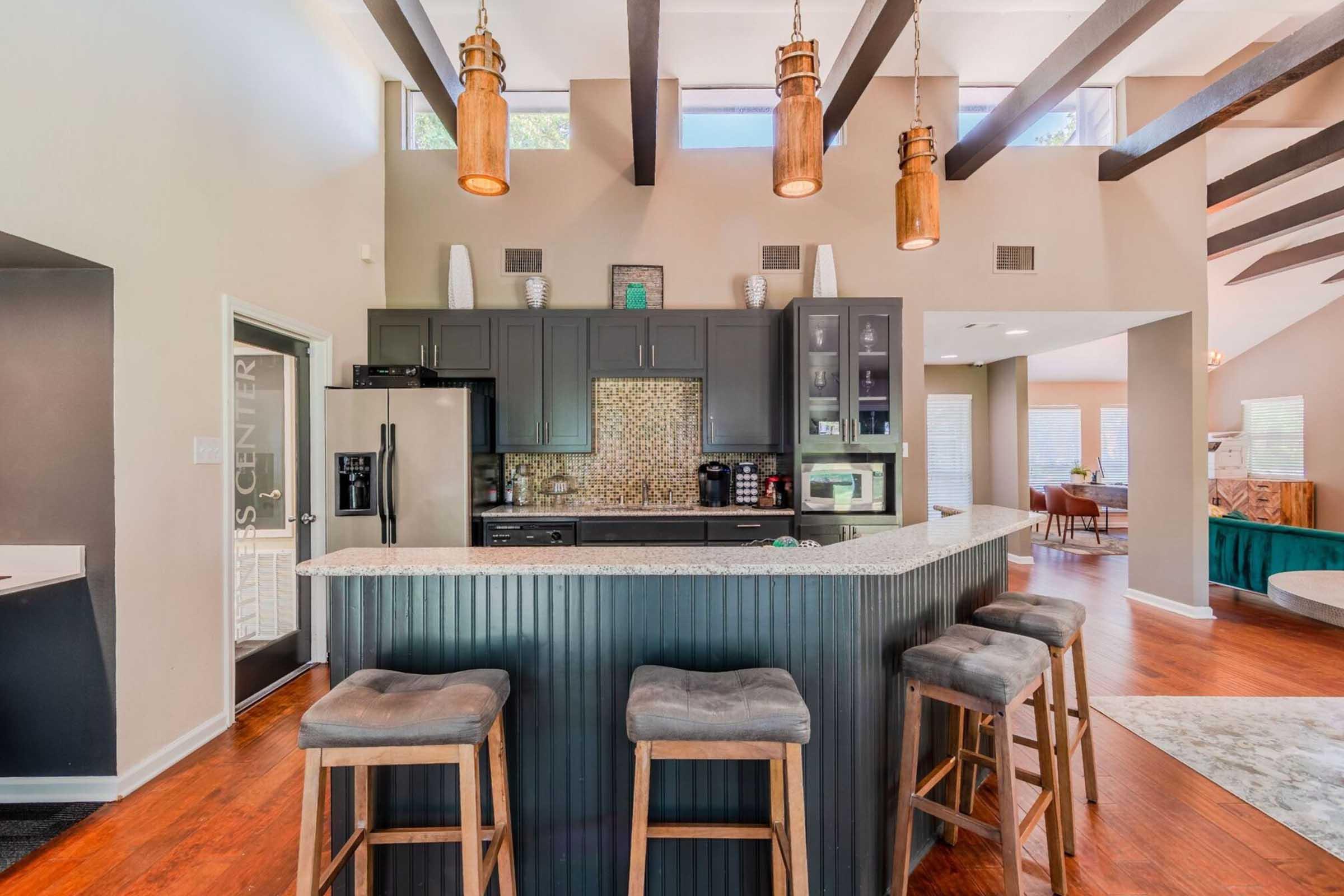 A modern kitchen with a central bar featuring three stools, dark cabinetry, and a mosaic backsplash. The space is brightened by pendant lighting and has wooden beams on the ceiling. There are large windows providing natural light, and the adjacent living area is visible in the background.