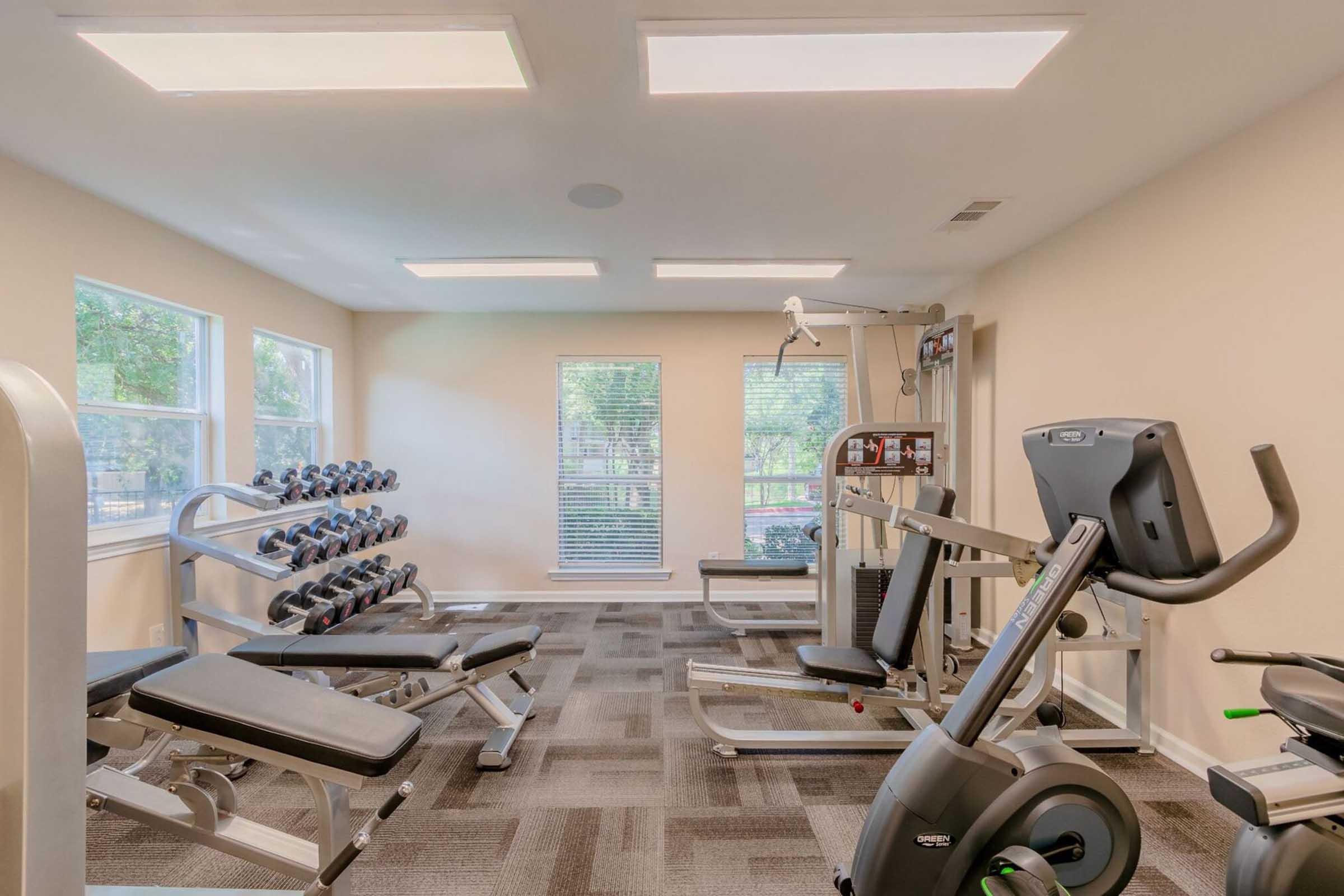 A bright and spacious gym featuring various exercise equipment, including dumbbells on racks, a weight bench, a multi-station machine, and a stationary bike. Large windows allow natural light to fill the room, enhancing the inviting ambiance.