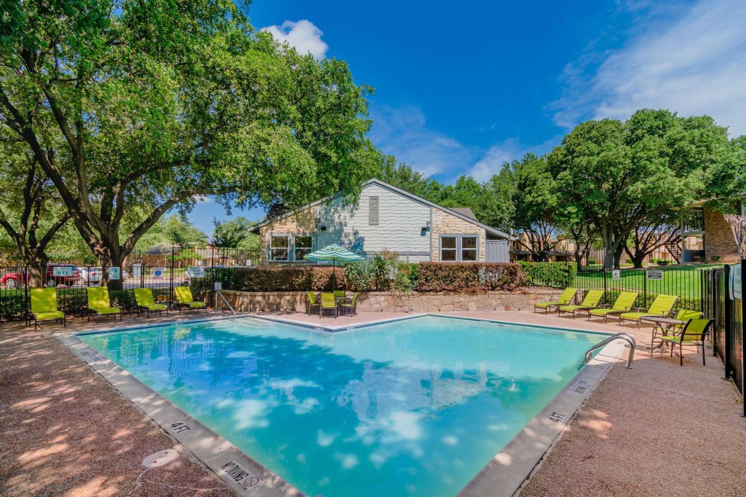 A clear blue swimming pool surrounded by lounge chairs and trees. In the background, a light-colored building and a well-maintained landscape. The sky is bright with a few clouds, creating a welcoming outdoor atmosphere.