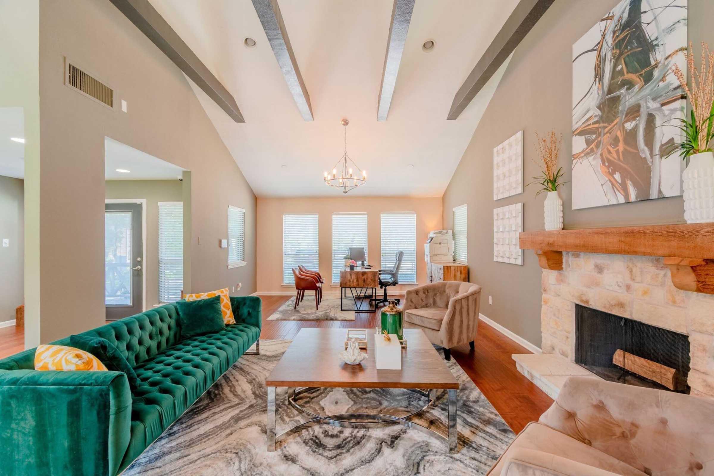 A modern living room featuring a green velvet sofa, a beige armchair, and a large coffee table on a patterned rug. The walls are painted a neutral color, with large windows letting in natural light. A chandelier hangs from the ceiling, and there are decorative pieces on the walls and around the fireplace.