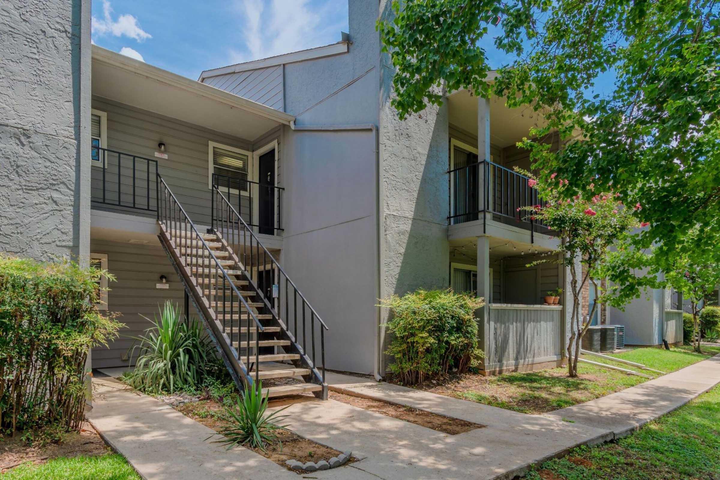 A view of two-story apartment buildings with a staircase leading to the upper level. The exterior features light gray walls and black railings. Lush greenery and bushes line the pathway, alongside neatly maintained grass and trees under a blue sky with scattered clouds.