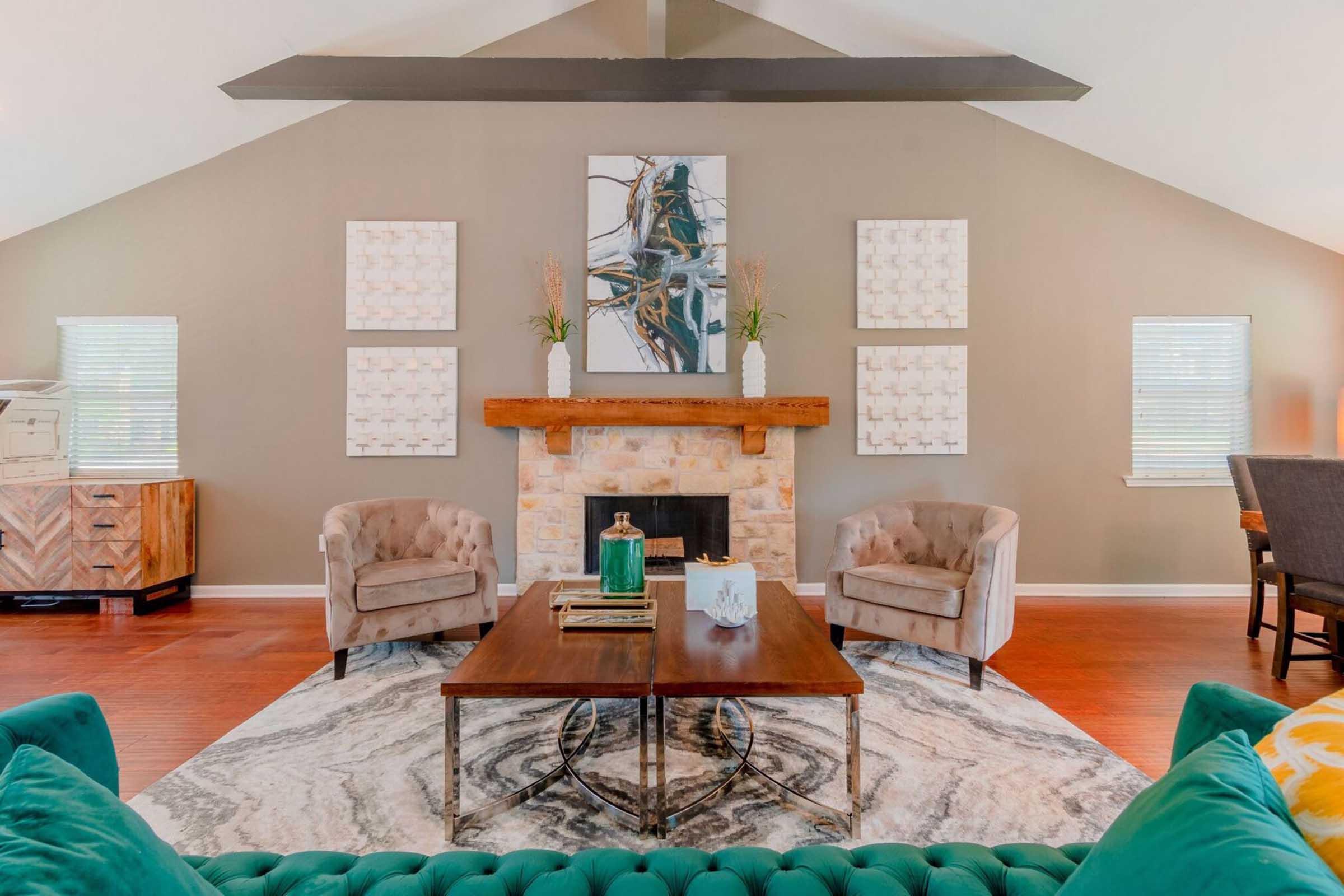 A spacious living room featuring a stone fireplace with a wooden mantel. Two upholstered chairs sit beside a wooden coffee table on a patterned rug. Simple wall art is displayed above the fireplace, and natural light enters through the windows. Decorative elements add a cozy touch to the modern decor.