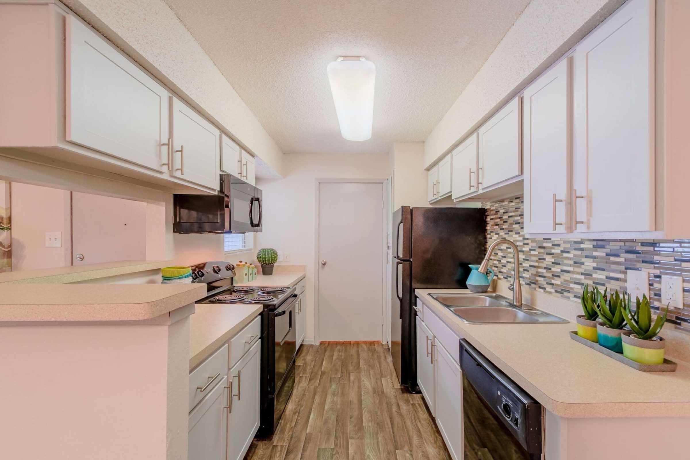 A modern kitchen featuring white cabinetry, a dark countertop, stainless steel appliances including a microwave and dishwasher, and a tiled backsplash. The space has laminate flooring and is well-lit by a ceiling fixture, creating a bright and open atmosphere. Green plants are placed on the counter for decoration.