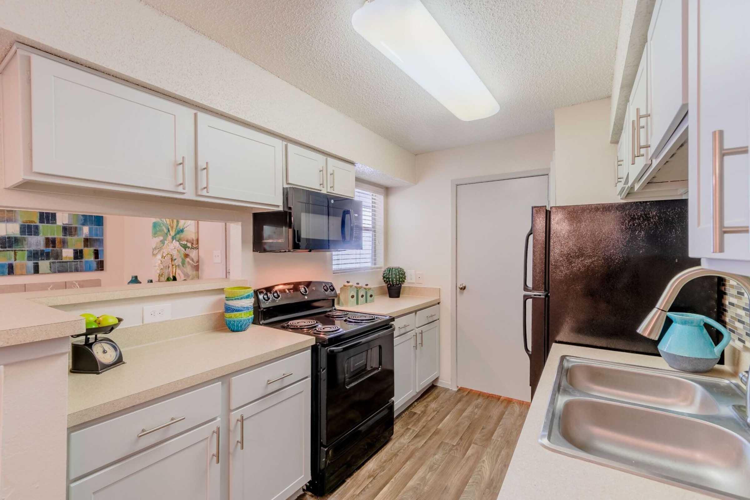 A modern kitchen featuring light-colored cabinets, a black stove, and a microwave. The countertops are beige with a dual sink. A dark refrigerator stands against the wall, and there's decorative wall art visible in the background. Natural light filters in through a small window.
