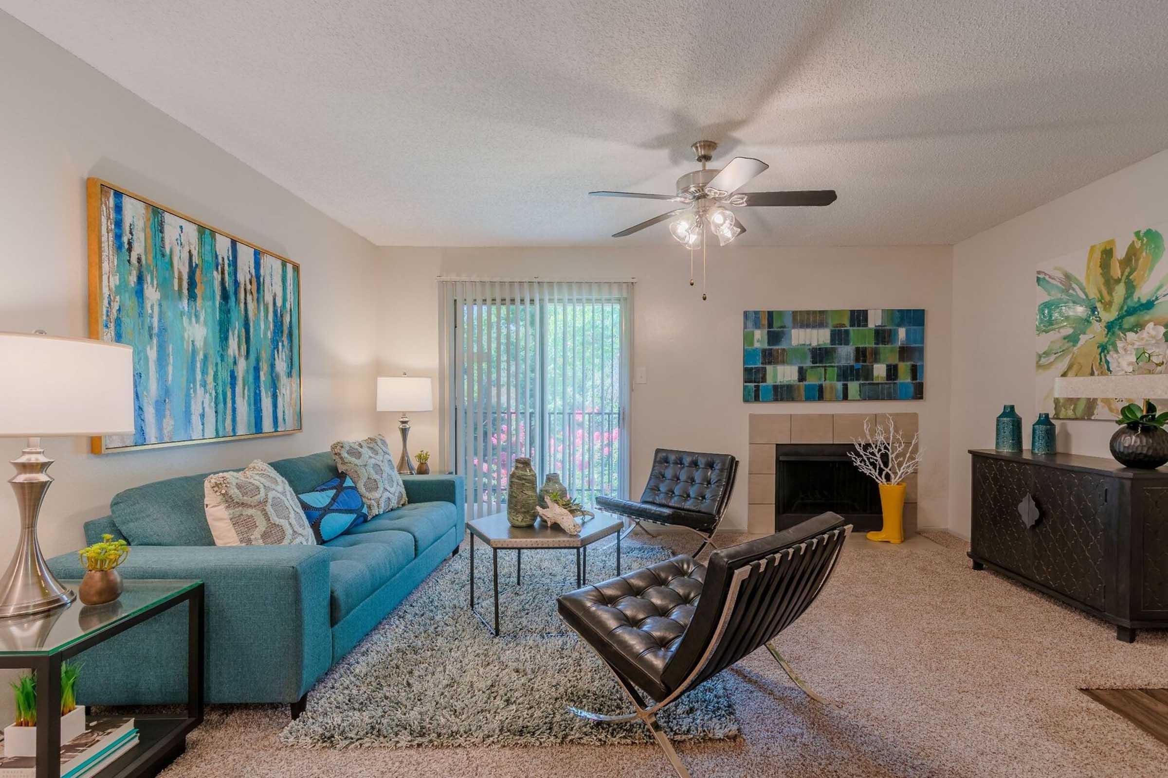 A modern living room featuring a light blue sofa with decorative pillows, two black leather chairs, and a glass coffee table. The room has art on the walls, a ceiling fan, and a fireplace. Large sliding glass doors lead to an outdoor space, allowing natural light to fill the area.