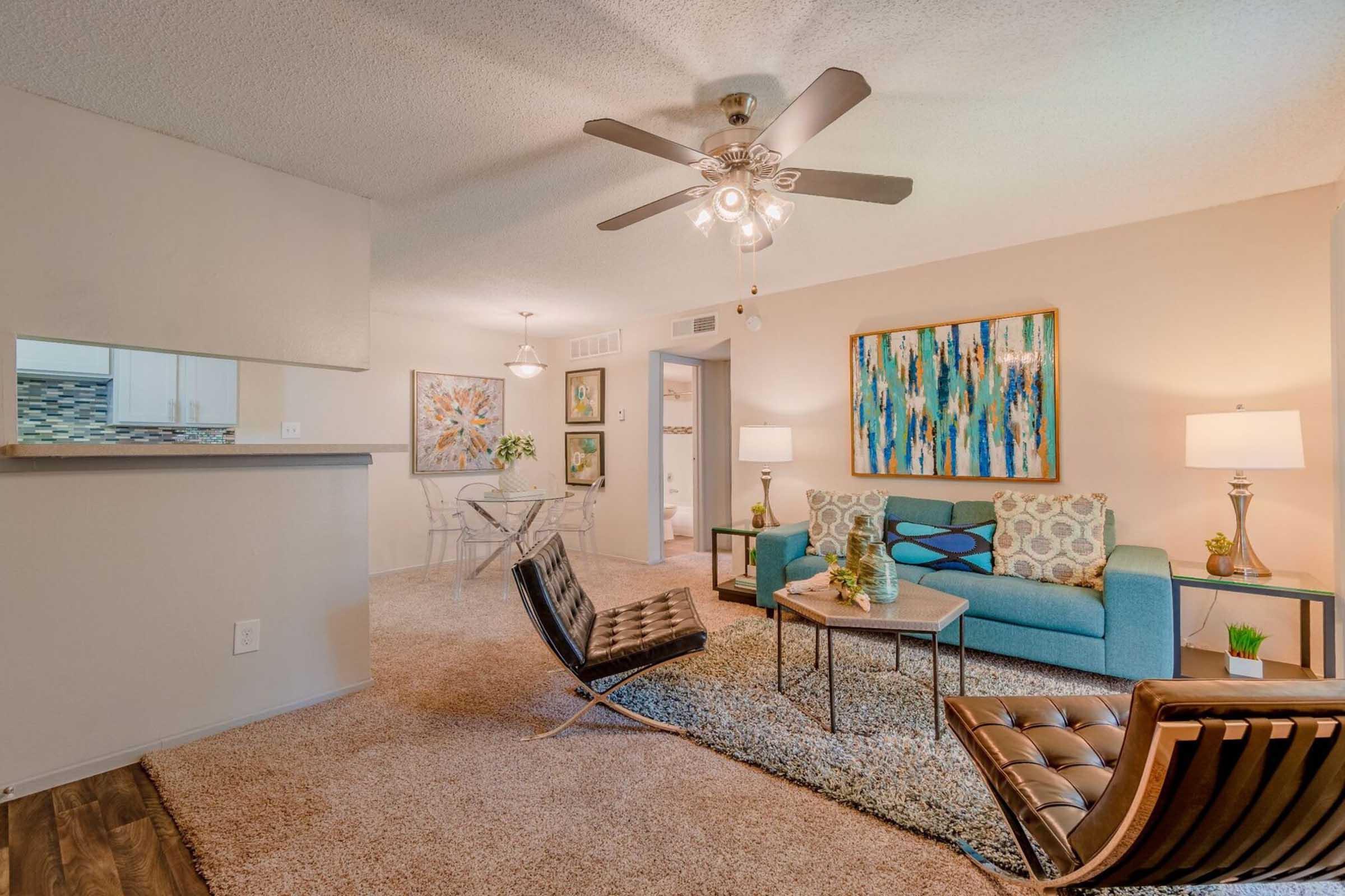 A stylish living room featuring a turquoise sofa, modern armchairs, and a decorative rug. The space is illuminated by a ceiling fan and floor lamps. Wall art adds color, while a glass dining table enhances the open layout. Soft, neutral tones create a cozy atmosphere.