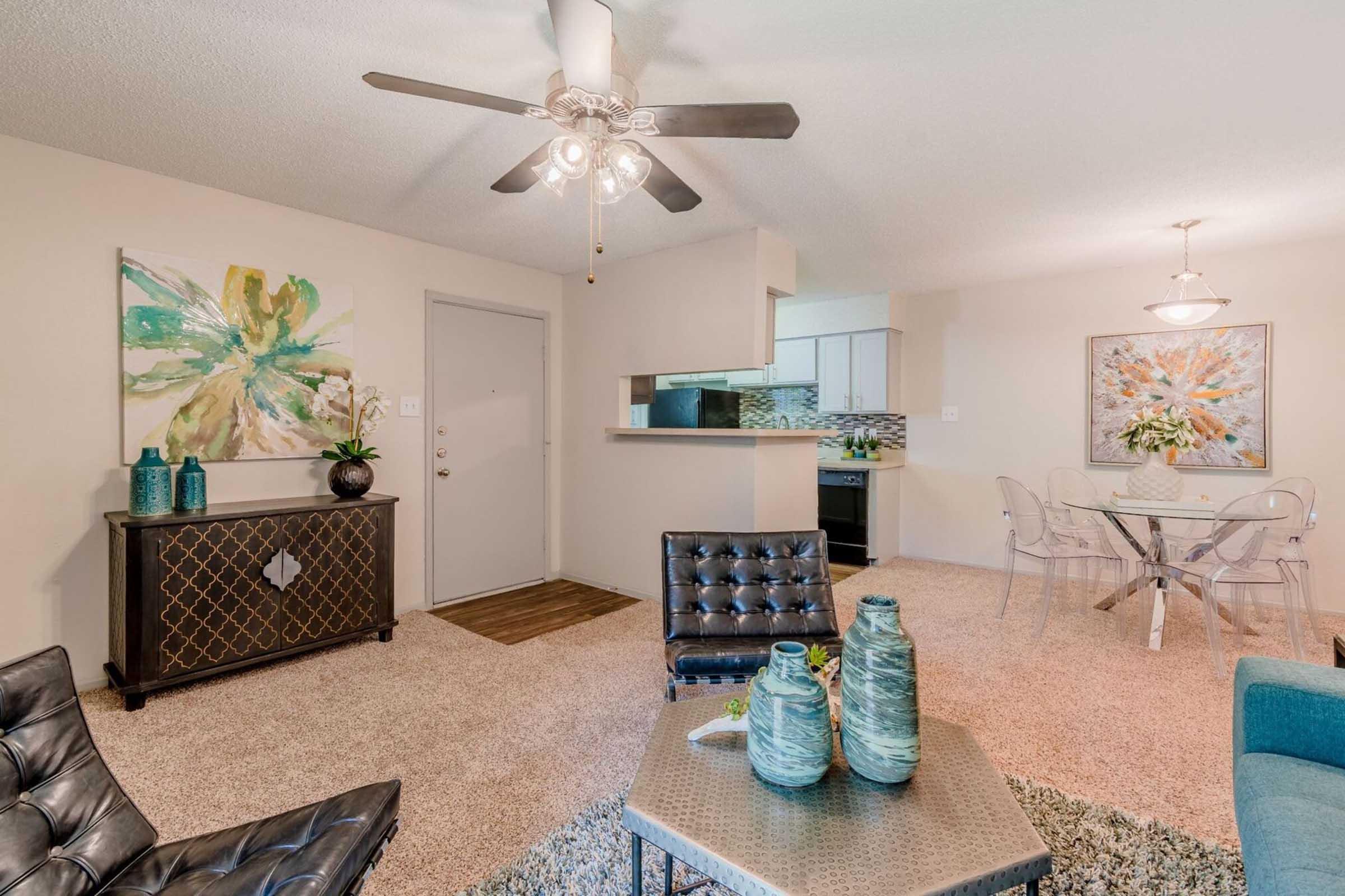 A cozy living space featuring a ceiling fan, a black leather couch, and a stylish coffee table with decorative vases. A modern dining area with clear chairs and an elegant table is visible. The walls are adorned with colorful abstract artwork, creating a welcoming atmosphere. The space has neutral tones and plush carpeting.