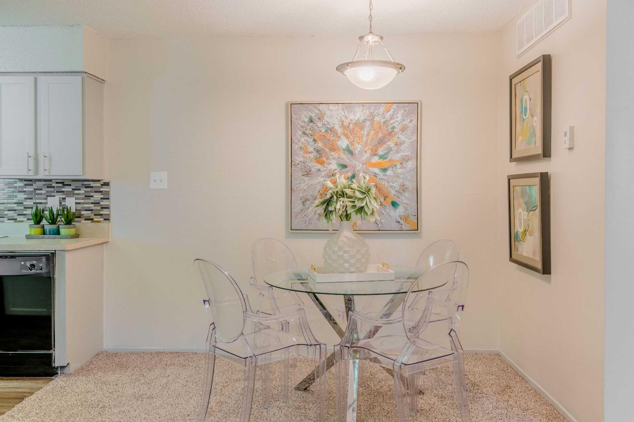 A stylish dining area featuring a round glass table surrounded by clear acrylic chairs. Above the table, a pendant light hangs, illuminating a large colorful abstract painting. Two framed artworks are on the wall nearby, and the adjacent kitchen area shows modern cabinetry and a decorative plant.