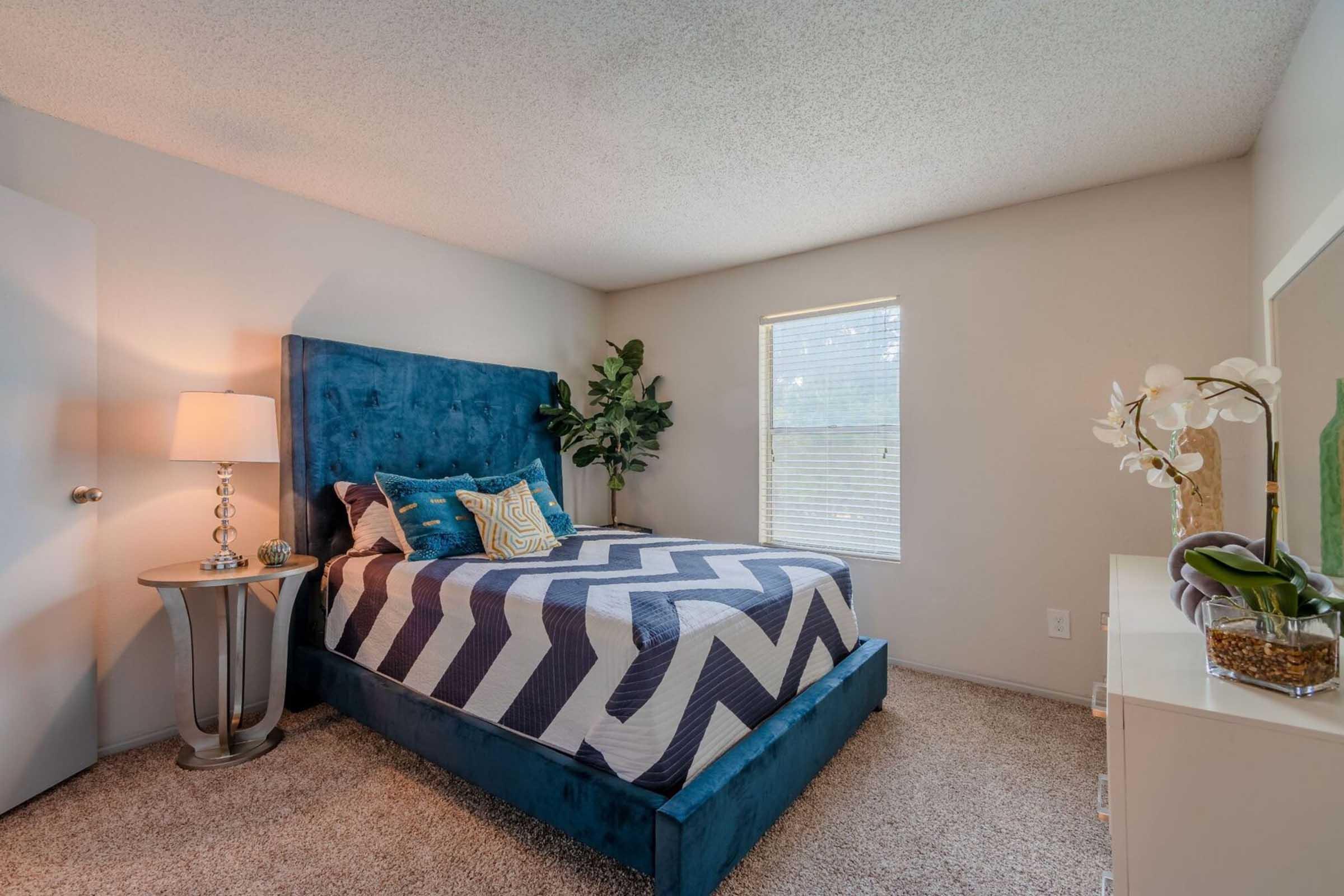 Cozy bedroom featuring a blue upholstered bed with a chevron-patterned duvet, a stylish nightstand with a lamp, and a window with natural light. Decorative plants and a white dresser add a touch of elegance to the warm, inviting space. Carpet flooring completes the room's comfortable atmosphere.