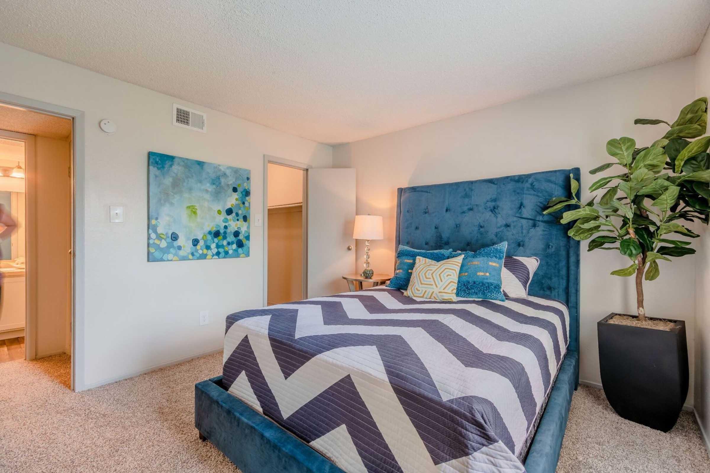A cozy bedroom featuring a blue upholstered bed with a zigzag patterned blanket. There's a plant in a black pot beside the bed and abstract artwork on the wall. A lamp provides soft lighting, and a doorway leads to a bathroom. The room has light-colored walls and carpet flooring.