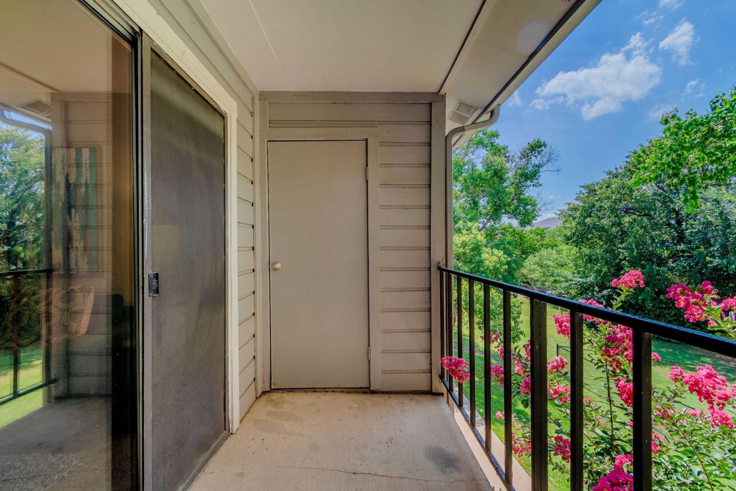 A balcony featuring a door leading inside, surrounded by greenery and bright pink flowers. The scene is well-lit with blue skies and fluffy clouds in the background, creating a cheerful and inviting atmosphere.