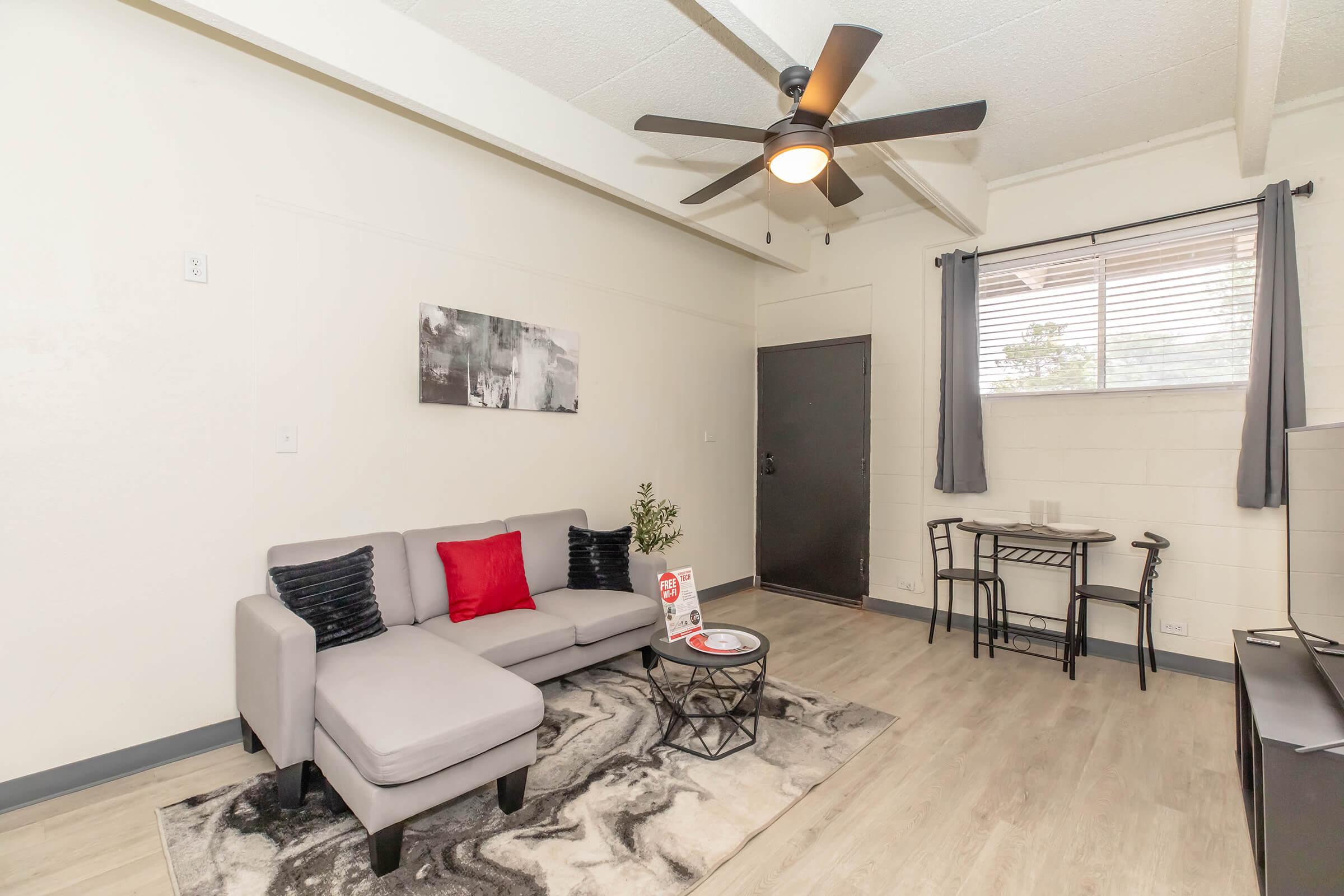 A cozy living room featuring a light gray sectional sofa with red accent pillows, a round coffee table, and a small dining table with two chairs. The space has a ceiling fan, a large window with gray curtains, and modern decor on the walls. The flooring is light wood, creating a warm and inviting atmosphere.