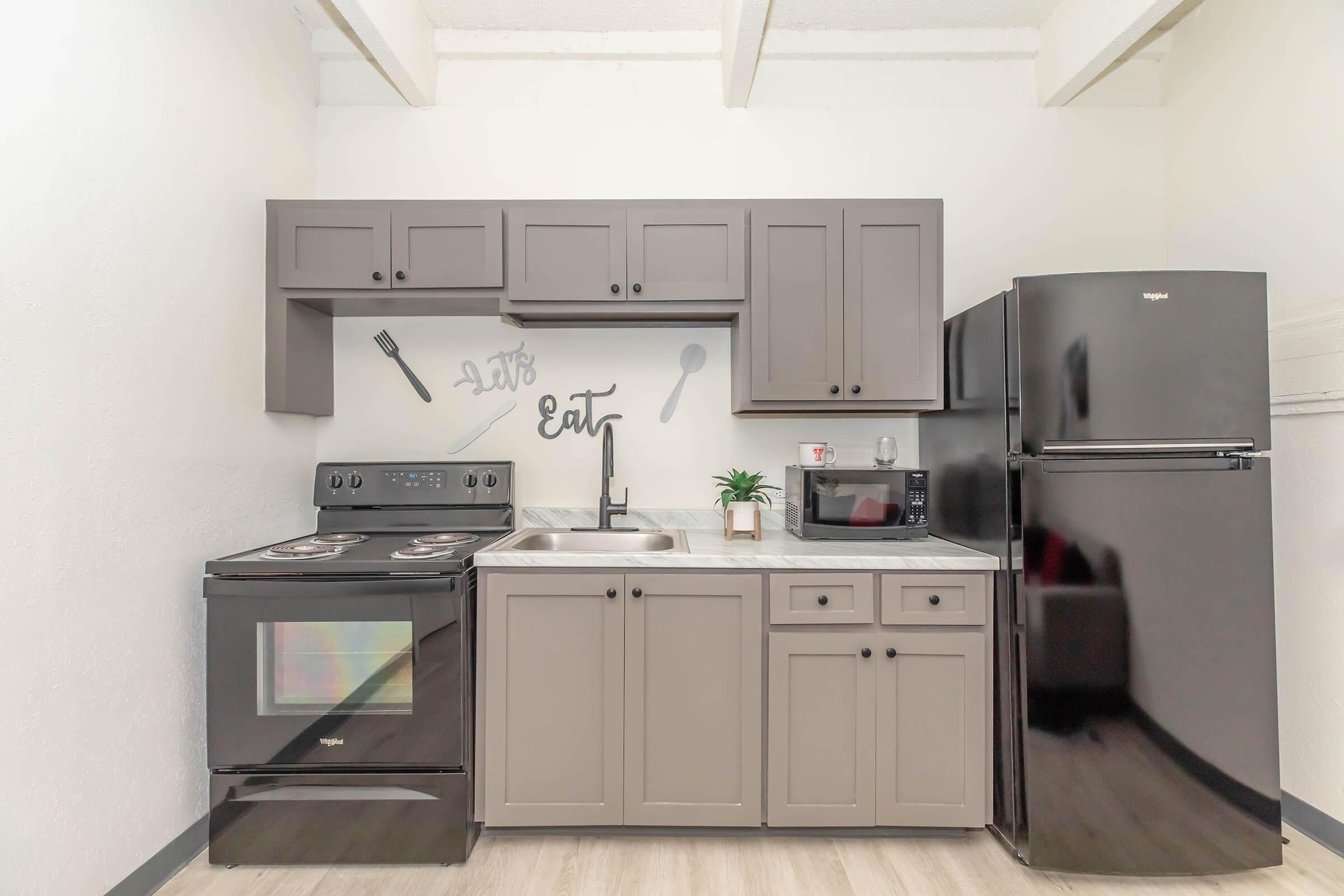 A modern kitchen with dark cabinetry, a black stove, and a matching refrigerator. The countertop is light-colored with a sink and a small microwave. The wall features a decorative "Let's Eat" phrase and kitchen utensil graphics, creating a cozy and inviting atmosphere.